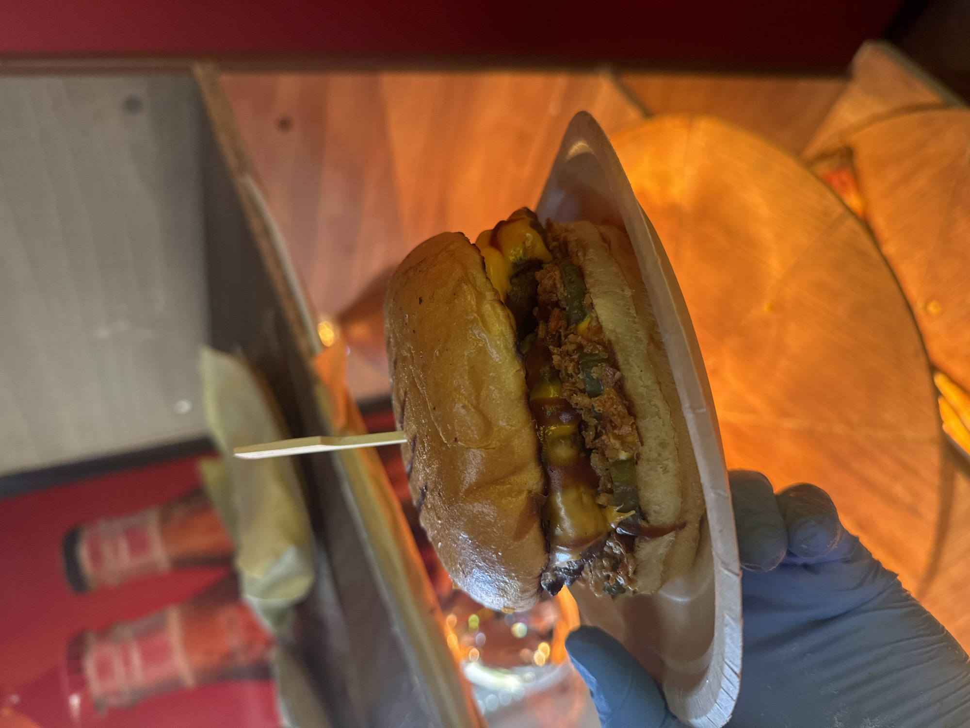 A close-up shot shows a person wearing a light blue glove holding a paper plate with a burger. The burger has a toasted bun, melted cheese, pickles, and barbecue sauce. In the background, out of focus, are two red-capped bottles on a shelf. The scene appears to be indoors, possibly in a restaurant or food stall, with warm lighting. The burger is the primary subject, centered in the frame, and the hand holding the plate is positioned in the lower-left corner. The overall aesthetic suggests a casual dining experience. The location in Sofia, Bulgaria is not evident from the image.