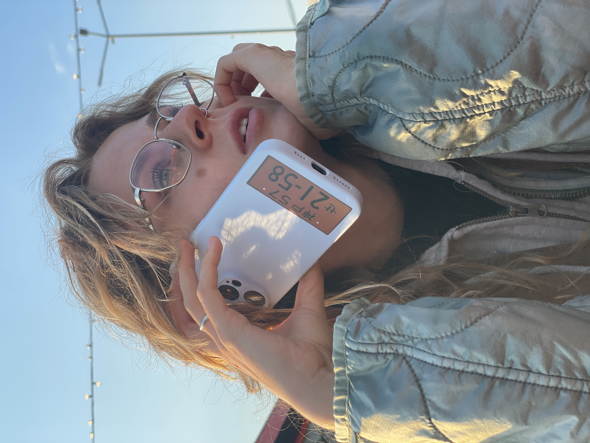A young woman with blond hair and glasses is holding a white smartphone to her ear, displaying the time "21:58" and some Japanese characters on the screen. She is wearing a gray quilted jacket and is likely outdoors, with a clear blue sky in the background. In the upper left corner of the image, there are lights suspended on a string. The location cannot be determined from the image.