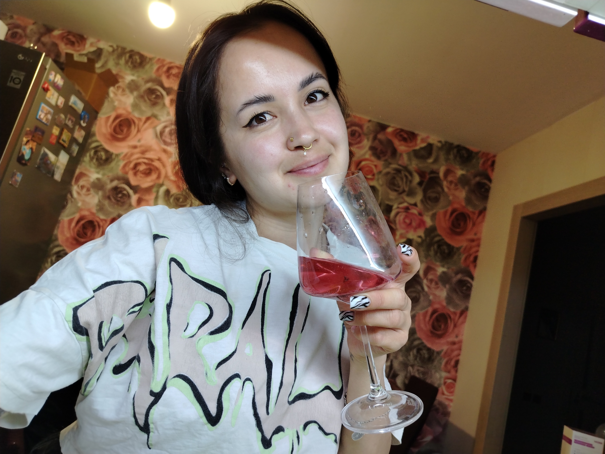 A woman is holding a wine glass filled with red wine. Her nails are painted with a black and white zebra pattern. She is wearing a white T-shirt with a black, light green, and beige design. She has a nose ring and earrings. The background is a wall covered in a floral pattern and a refrigerator with magnets on it. A doorway is visible in the background. The location is Noviy, Russia.