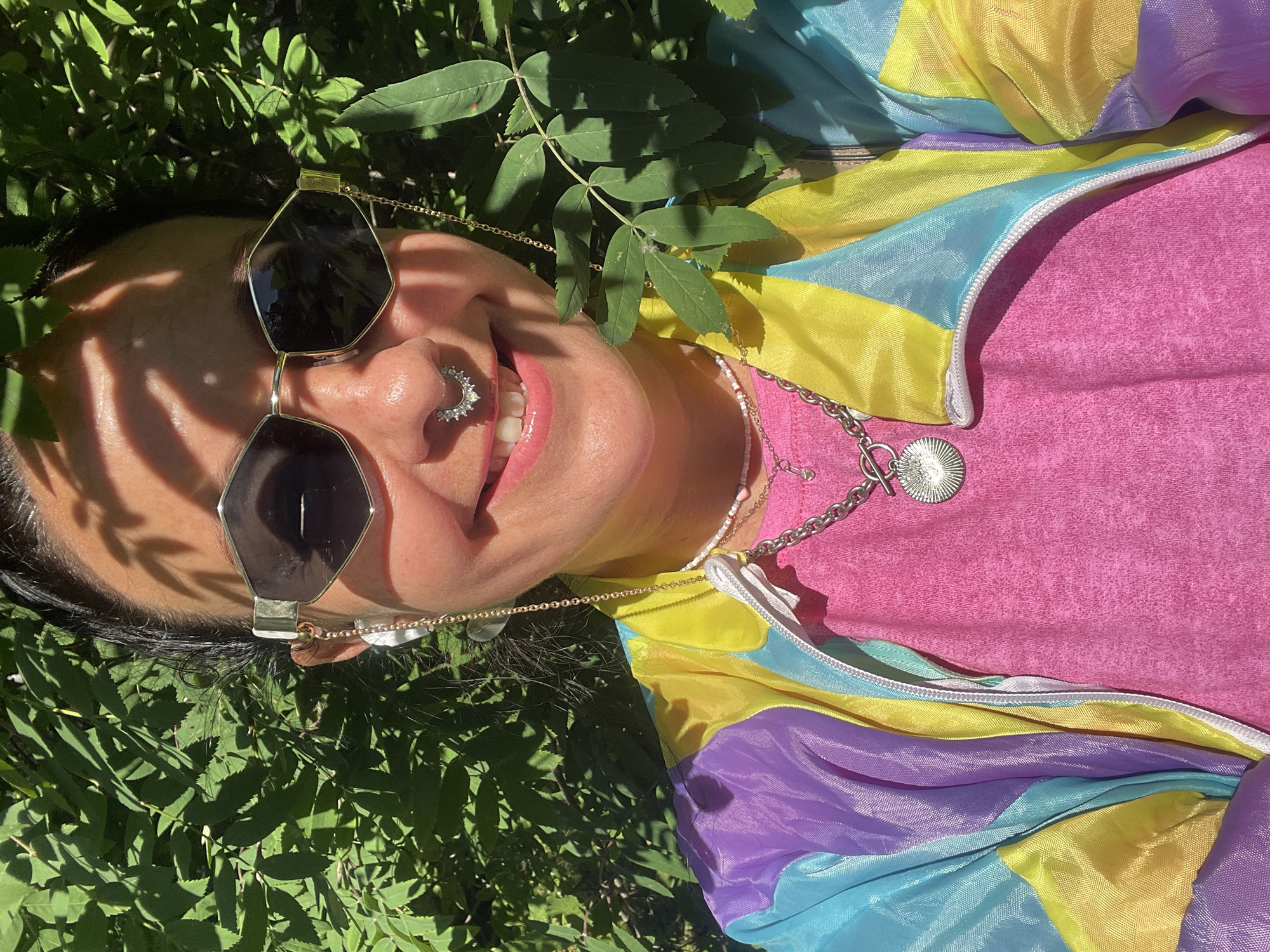 A close-up selfie of a woman with dark hair, wearing sunglasses, a septum piercing, and a colorful windbreaker over a pink t-shirt. She is outdoors, with green foliage in the background. The lighting suggests it is daytime. The image appears to have been taken in Saint Petersburg, Russia, though no specific landmarks are visible.