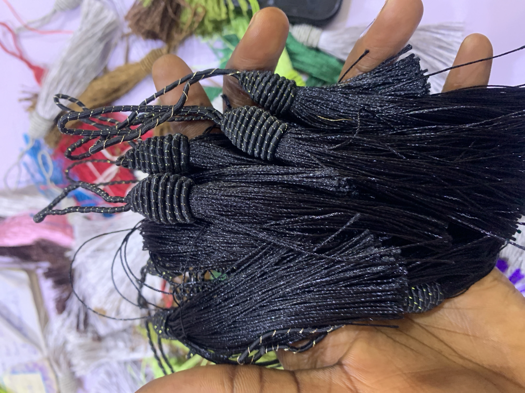 A person's hand, exhibiting medium-brown skin tone, is holding a cluster of black tassels. The tassels are adorned with thin, gold-colored threads that are twisted around the upper portion of each tassel, creating a ribbed effect. The tassels themselves are made of numerous fine, black strands that cascade downwards. In the background, a variety of other tassels and materials, in colors such as white, pink, brown, and green, are visible, suggesting a market or craft stall setting. The background is out of focus, with some areas appearing brightly lit and others in shadow. The overall impression is of a close-up view of decorative craft items being displayed or handled. It is not possible to determine the specific location in Gura Top, Nigeria from the visual information provided.
