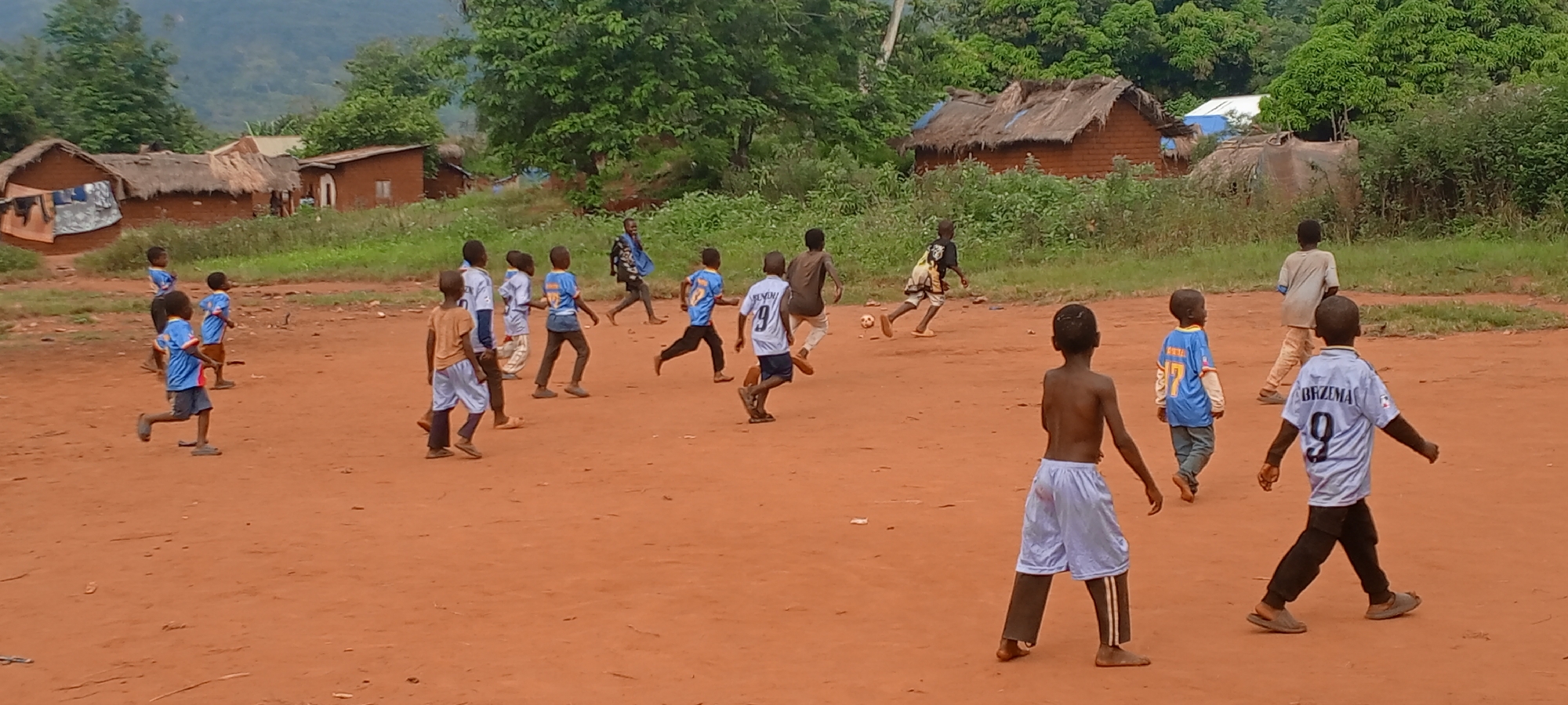 In Ngalula, Democratic Republic of the Congo, a group of children play soccer on a dirt field. Some wear soccer jerseys, one with "BRZEMA 9" printed on the back. The environment features simple buildings, some with thatched roofs, indicating a rural setting. Trees and vegetation are visible in the background.