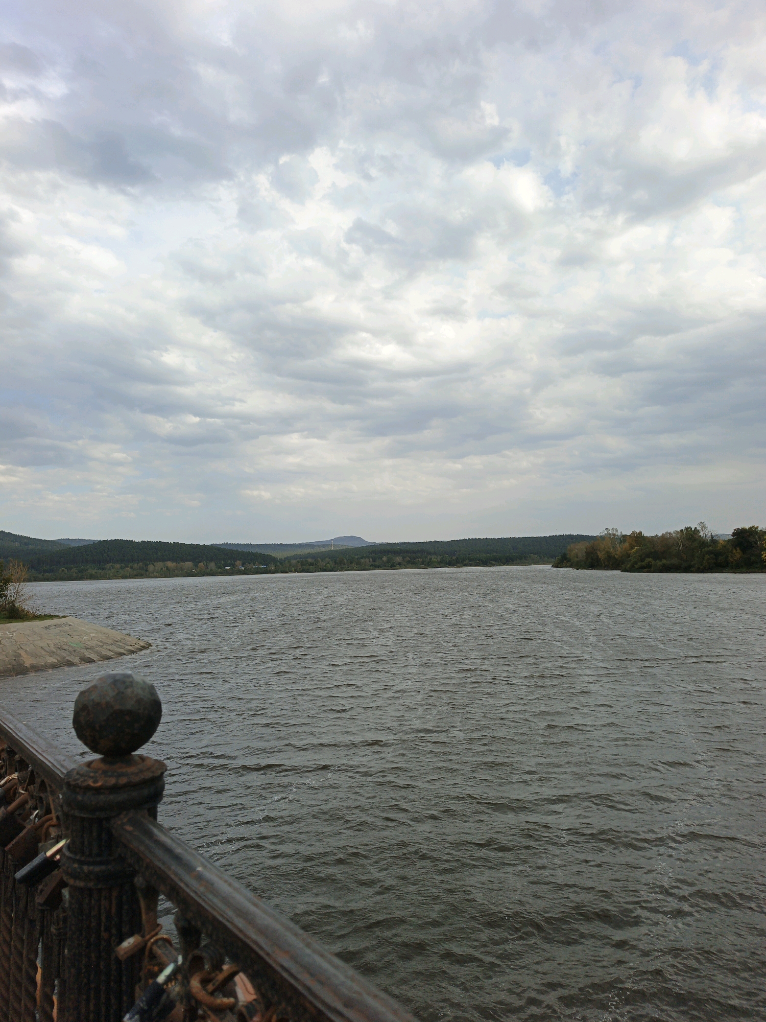 The image depicts a wide, choppy body of water, likely a reservoir or lake, under a cloudy sky. In the foreground, a rusty metal railing is visible, adorned with several padlocks, suggesting it is part of a bridge or promenade. The railing is ornate, with a decorative spherical finial at the top of a post. To the left, a concrete embankment slopes down to the water. The background features a dense, green treeline that stretches across the horizon, with rolling hills and mountains visible in the distance. The overall atmosphere is subdued, with muted colors and diffused light typical of an overcast day. This scene is likely located on the Zlatoust Reservoir in Zlatoust, Russia, a prominent landmark known for its scenic views and recreational activities. There are no people visible in the image, and the focus is on the natural landscape and the man-made structure of the railing.