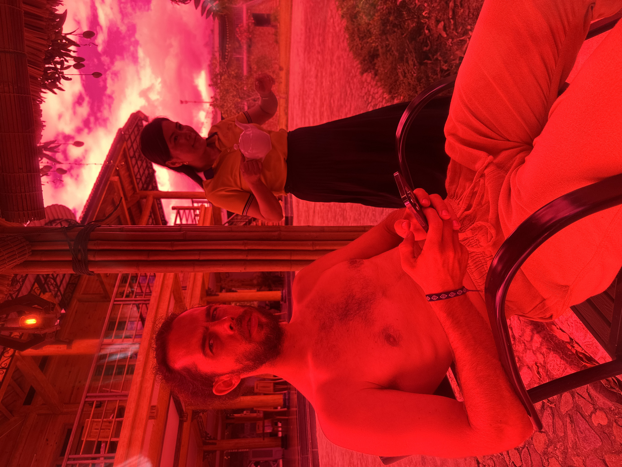 The image, taken in Nghia Lo Ward, Vietnam, is characterized by a strong red/pink monochromatic tint. In the foreground, a shirtless Caucasian male with a beard and dark hair is seated in a dark-framed chair. He wears light-colored trousers and a patterned bracelet on his left wrist, holding a dark smartphone in his right hand. His gaze is directed towards the viewer. Behind him and to the right, an East Asian female stands, wearing an orange polo shirt with dark trim and a long dark skirt. She holds a light-colored teapot with both hands. The scene is set under a thatched roof supported by dark bamboo poles. In the background, a multi-story building with balconies and railings is visible to the left, while another structure and dense vegetation appear to the right, beneath a heavily pink and cloudy sky. A red light fixture hangs from an overhead beam. The ground transitions from patterned paving stones in the foreground to natural terrain.