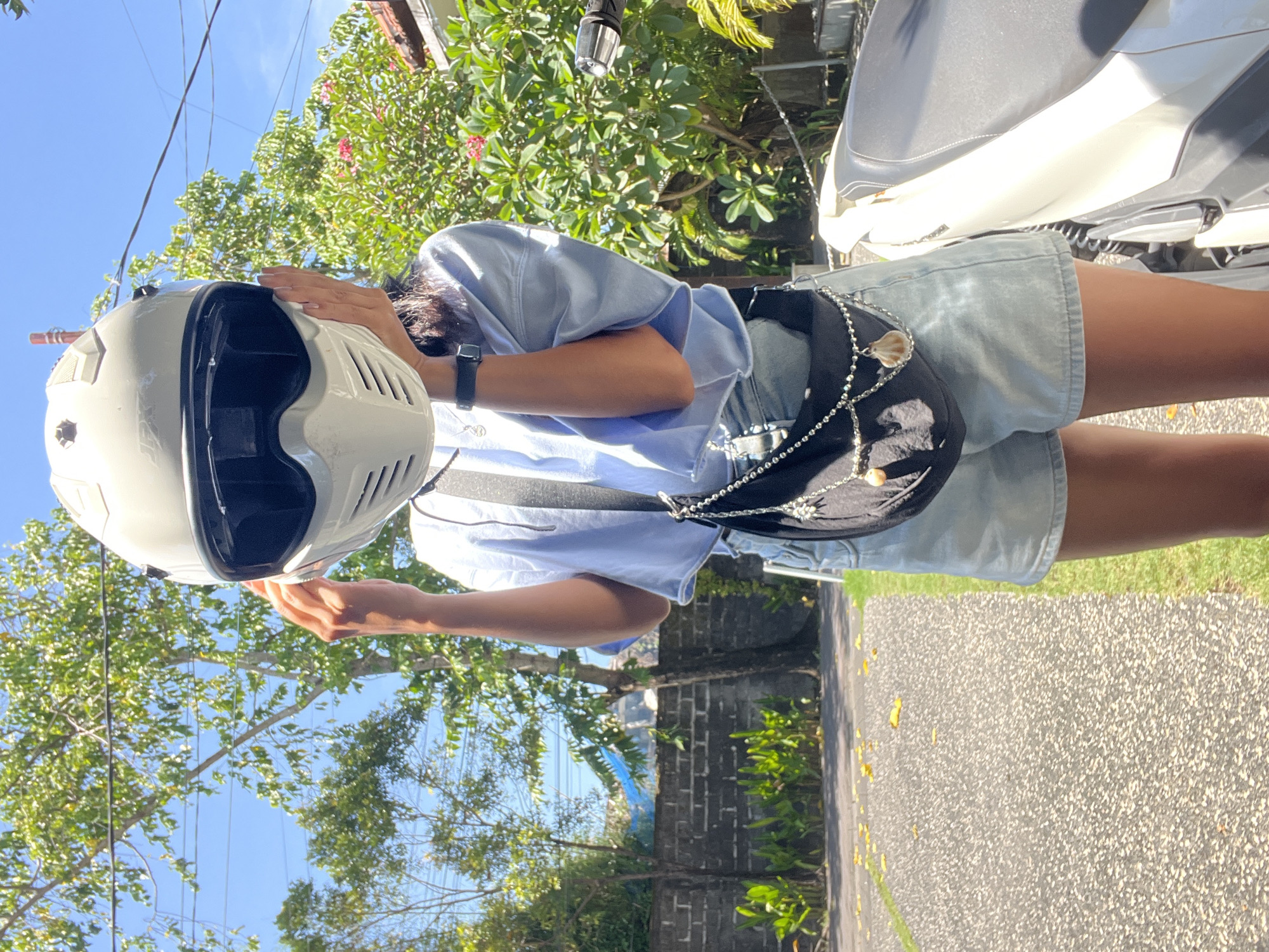The image shows an individual in Kuta Selatan, Indonesia, standing outdoors on a paved surface next to a parked white scooter or motorcycle. They are wearing light blue denim shorts and a light blue top, with a black crossbody bag adorned with a silver chain and seashell charms. The person holds a white motorcycle helmet with a black visor in front of their face with their right hand. Their left hand is raised towards their face. A watch is visible on their left wrist. Lush green foliage and trees are visible in the background, partially obscuring a brick wall and a blue tarp. Overhead, power lines crisscross the sky. The lighting indicates it is daytime.