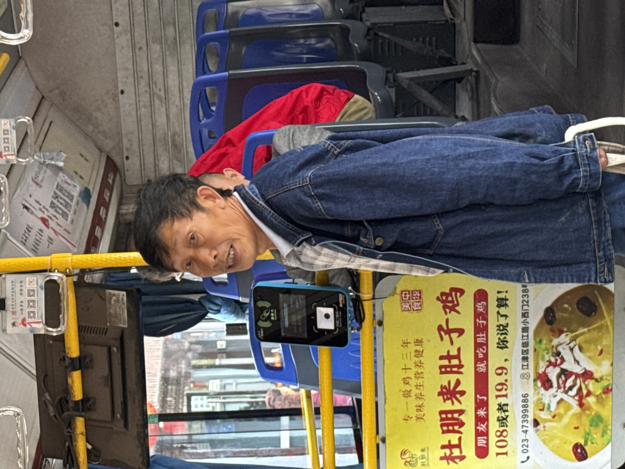 A middle-aged Asian man with short, graying hair and wearing a blue jean jacket and a checkered shirt underneath sits on a bus in Jiangjin, China. He is facing the camera with a slight smile. Behind him are blue bus seats and a red jacket is draped over one of them. Adjacent to him is a yellow pole with a digital display showing bus information. An advertisement for "Du Peng Lai" chicken soup is attached to the pole, indicating a local specialty. The advertisement includes text in Chinese, a phone number (023-47399886), and a partial address (江津区临江路小西门238). Through the bus windows, blurred buildings and streets can be seen, suggesting the bus is in motion.