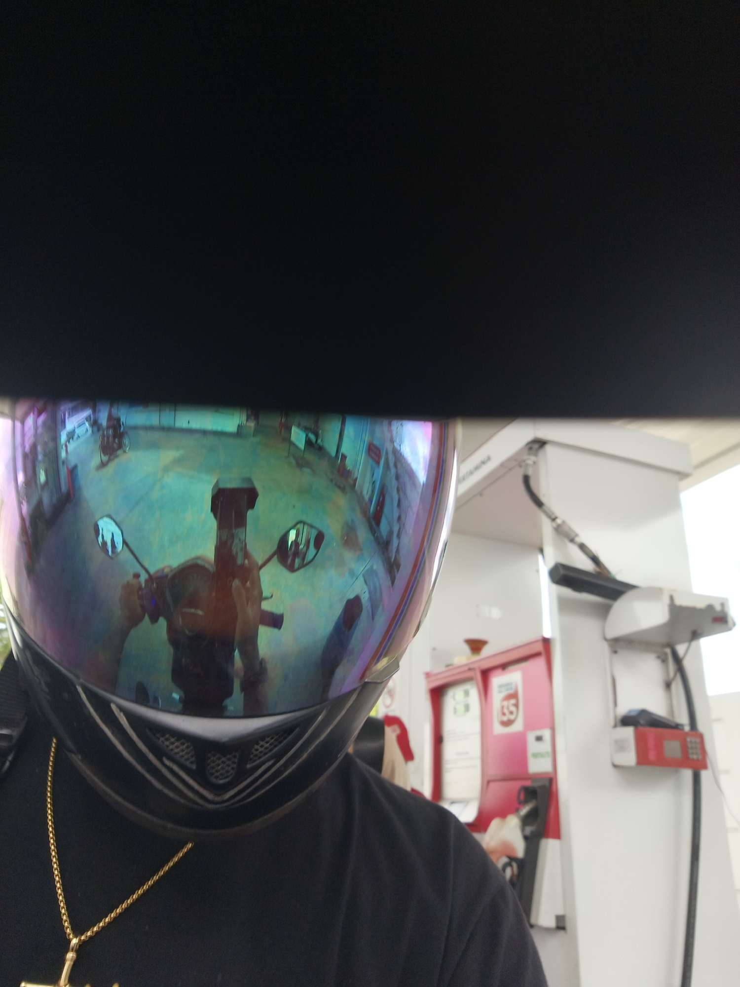 The image is a selfie taken by someone wearing a black t-shirt and a motorcycle helmet with a reflective visor. The visor reflects the surrounding environment, which appears to be a gas station. In the reflection, a person is seen holding a smartphone to take the selfie, and other motorcycles and a gas pump are visible. The gas pump has a red body and a white top, with some labels and a nozzle. The ground is concrete, and there are some buildings or structures in the background. The location is identified as a gas station in Denpasar, Indonesia.