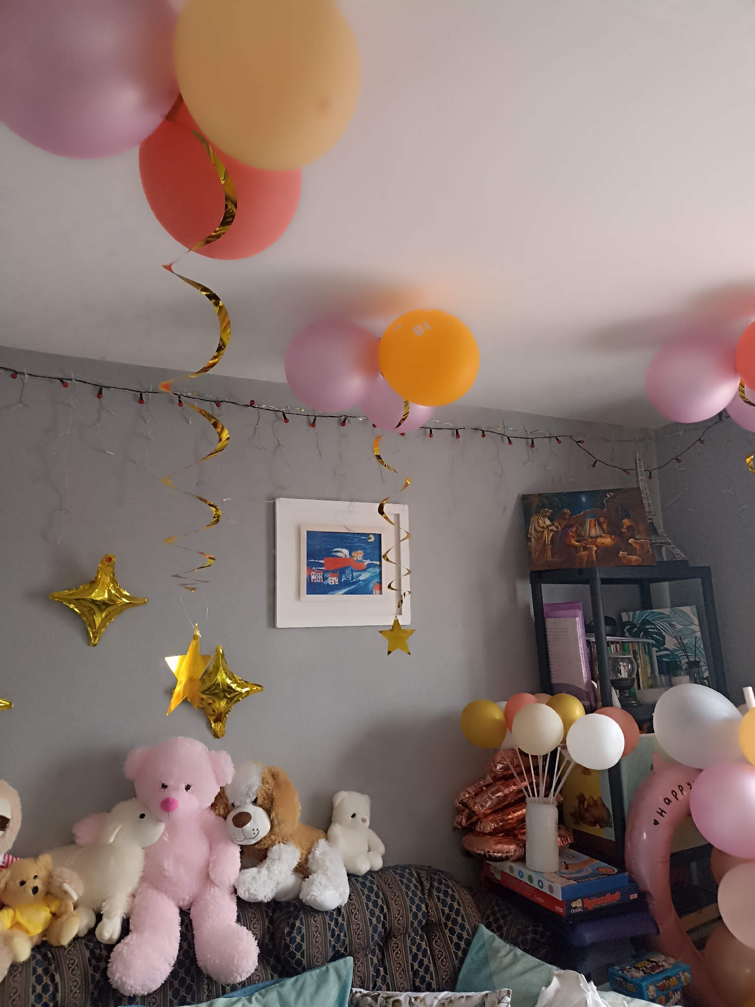 The image depicts a decorated room, likely for a celebration, with an assortment of balloons and stuffed animals. Pink, orange, and yellow balloons are scattered across the ceiling and clustered on the right side of the frame. Several gold star-shaped balloons are affixed to the grey wall. A string of small red lights is also visible along the wall. On the wall, there are two framed pictures. The stuffed animals are arranged on a couch in the foreground, including a large pink teddy bear, a brown and white dog, and several smaller bears and bunnies. Behind the couch, on shelves, are books and decorative items, including a framed nativity scene. The overall impression is that of a festive, indoor setting, with no individuals present. The location of Annagry, Ireland, is not visually ascertainable from the image.