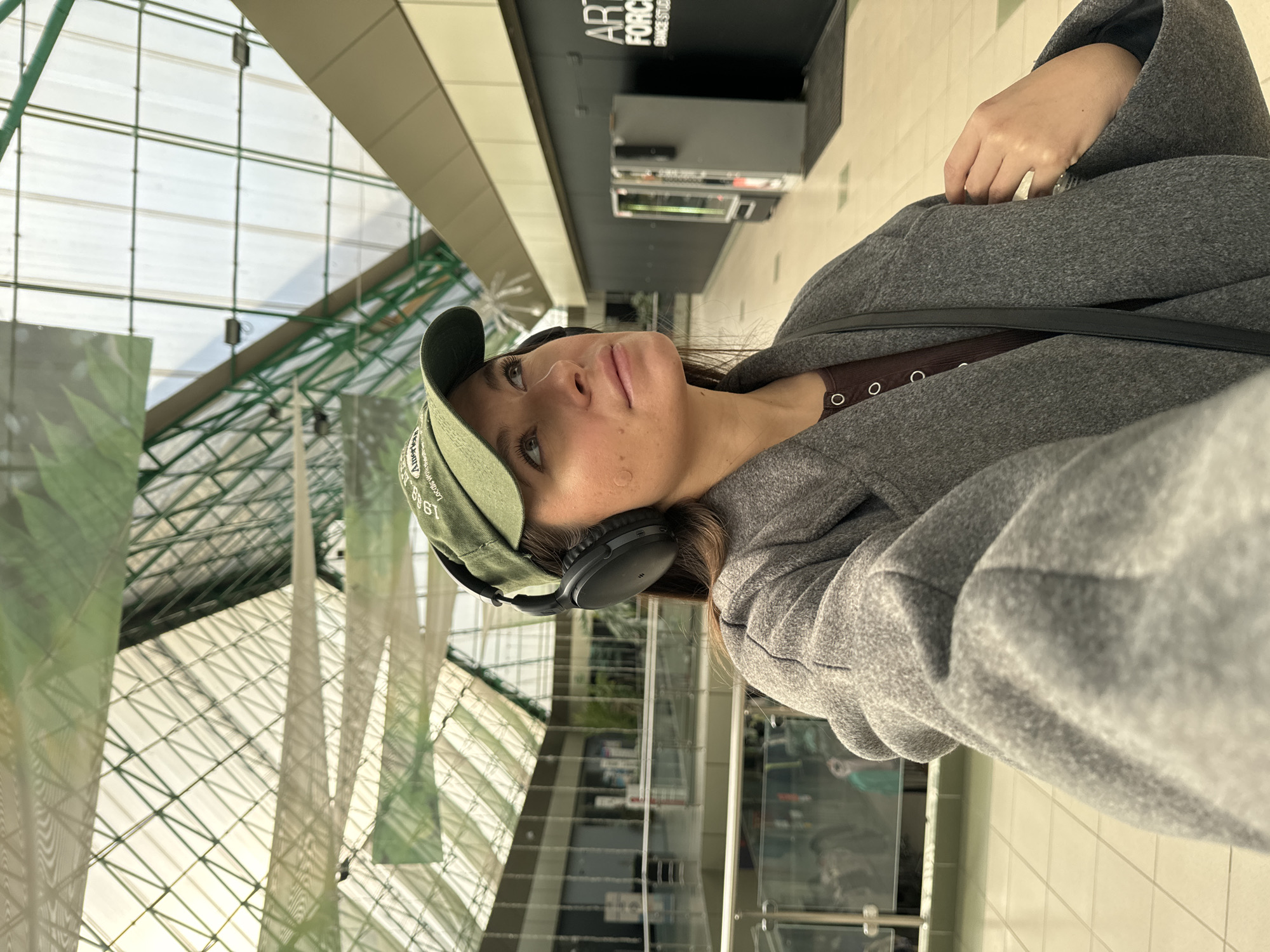 A female subject is depicted in a selfie within the common area of the TC Kurs shopping mall, located at Mozhaiskoye Shosse, 71, Odintsovo, Russia. She wears an olive green baseball cap with "American" visible text, black over-ear headphones, a brown ribbed long-sleeve top, and a grey woolen-texture coat. A light-colored circular patch is present on her left cheek. Her gaze is directed upwards and slightly to her right, and her right hand holds the lapel of her coat. The background reveals a multi-level interior with a prominent green-framed glass skylight roof forming a geometric grid pattern. To the subject's right, a black wall displays "ART FORCE DANCE STUDIO" in white lettering, accompanied by two vending machines (one light grey, one dark grey/black) and a black floor mat. Glass and metal railings delineate a lower level, and the floor is surfaced with light-colored tiles.