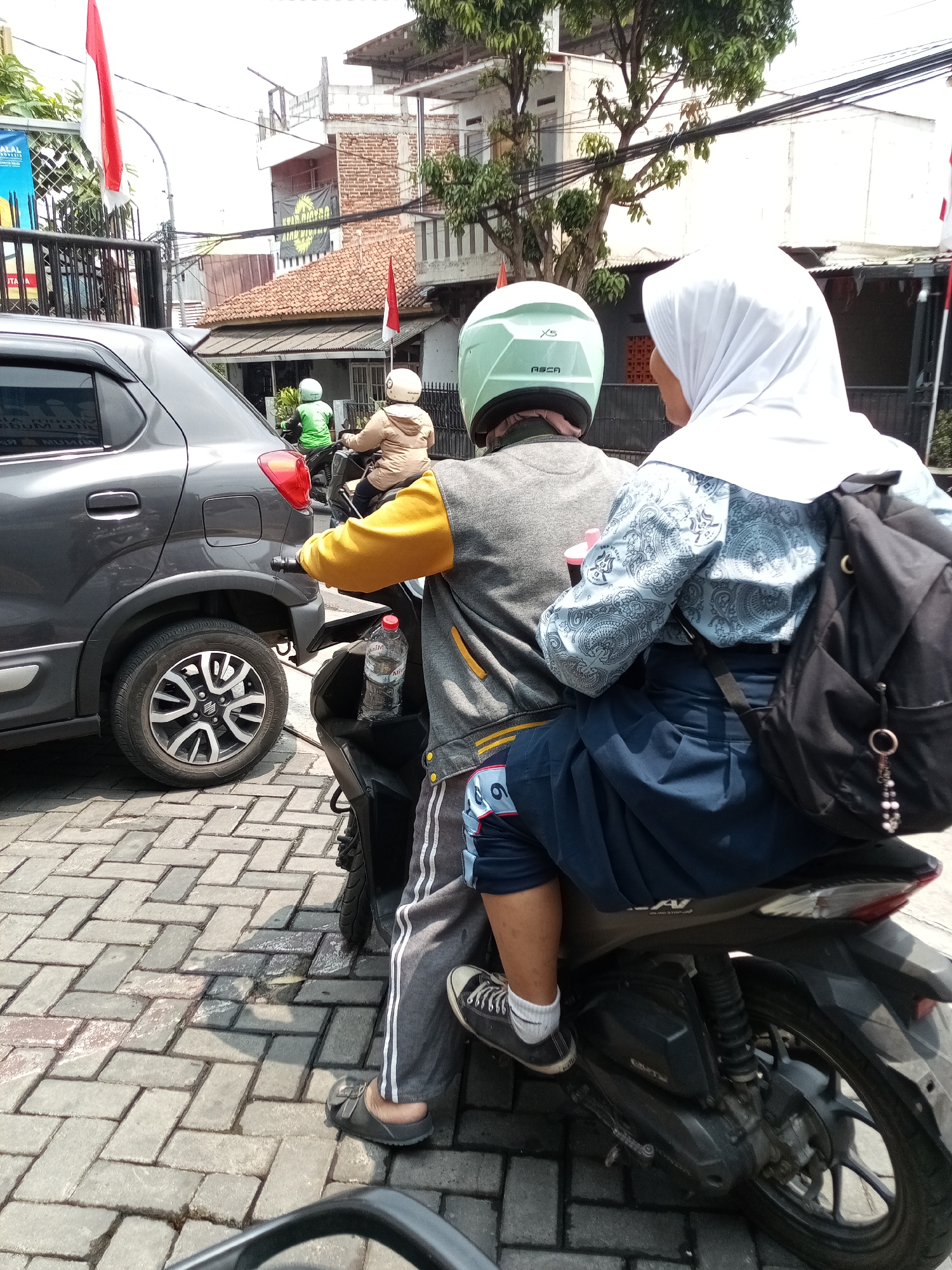 Two individuals are riding on a motorcycle in what appears to be an urban environment, possibly in Cimahi, Indonesia. The person in the rear is wearing a white hijab and a patterned blue top, with a black backpack slung over their shoulder. The person in the front is wearing a light green helmet and a yellow and gray jacket. They are on a street with a brick-paved surface, and a gray car is parked to the left of the motorcycle. Red and white Indonesian flags are visible in the background, suggesting a national holiday or celebration. The scene is captured in daylight, with shadows indicating the sun is high in the sky. There are buildings and trees in the background, typical of a city landscape.