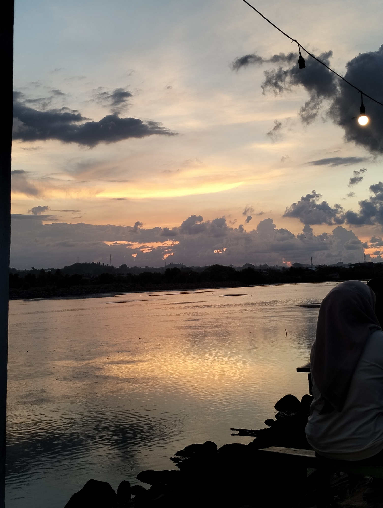 A low-angle, medium shot captures a sunset over a body of water, likely a river or lake, in Lhokseumawe, Indonesia. In the lower right foreground, a person wearing a hijab and white top sits with their back to the viewer on a bench or ledge overlooking the water. Rocks and silhouetted debris are scattered along the water's edge, adding depth to the scene.
The body of water reflects the warm colors of the setting sun, creating a shimmering effect. In the background, a dark treeline or low hills are visible, suggesting the outskirts of the city. Utility poles and towers stand out against the horizon.
Above, the sky is filled with a mix of dark, gray clouds and streaks of orange and yellow from the setting sun. A string of lights, typical of outdoor cafes or public spaces, is strung across the top of the frame. One light bulb is illuminated, casting a soft glow.
The photo is taken from a slightly obscured vantage point, possibly from within a building or under a shelter, as evidenced by a dark vertical element on the left edge of the frame. The overall atmosphere is calm and reflective, capturing a tranquil moment at sunset.