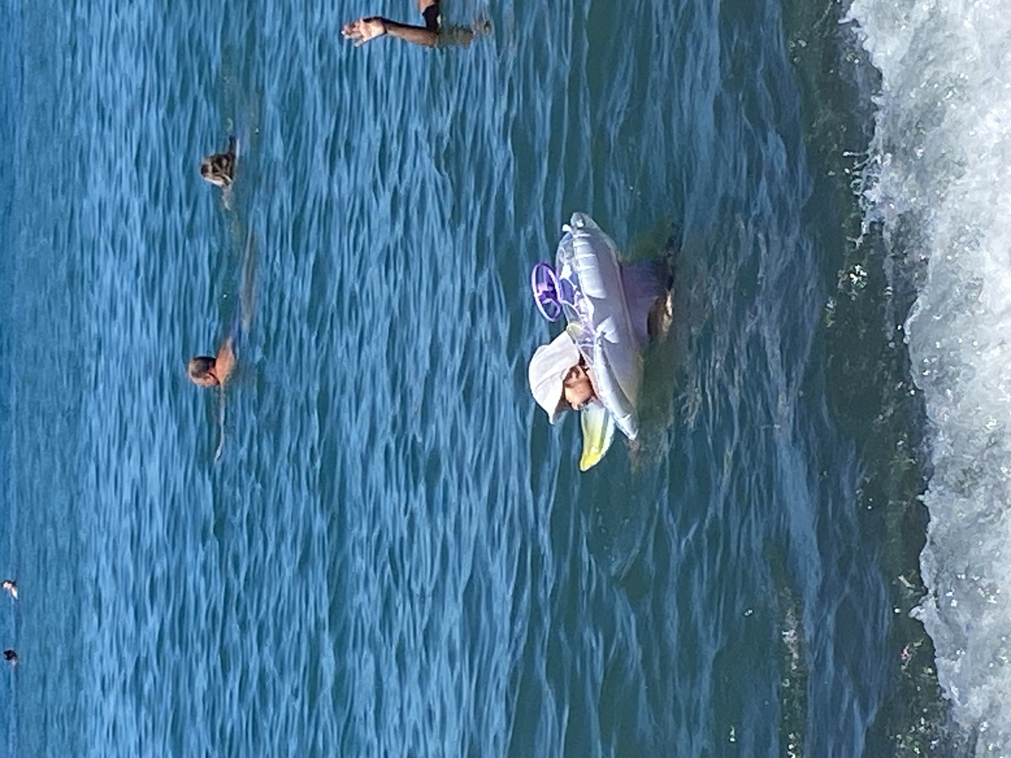 The image shows a shallow body of water, likely the Black Sea, near Sochi, Russia. In the foreground, a wave is breaking. In the middle ground, a baby wearing a sun hat is floating on an inflatable toy in the water. The baby is lying on its back and appears to be in a relaxed state. In the background, several adults are swimming in the water. One person is visible from the shoulders up, facing away from the camera. Another person is partially visible, also facing away. A third person's arm is raised, and they appear to be waving. The water is a deep blue, and the sky is not visible. The overall impression is a scene of a casual day at the beach.