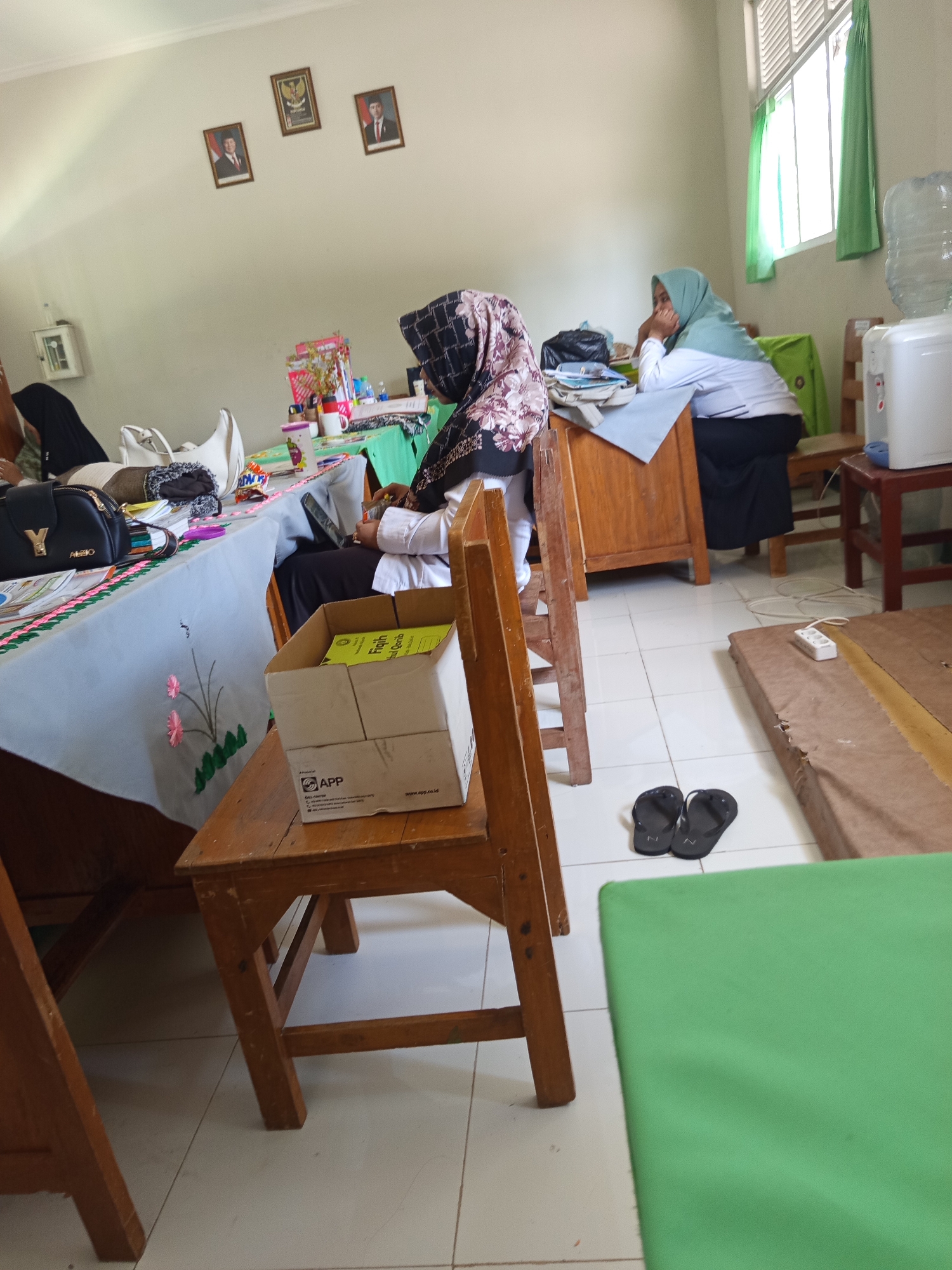 The image depicts an indoor scene, likely a classroom or common area, with three individuals present. One person, wearing a black hijab with a floral pattern, is seated at a table covered with a light blue tablecloth adorned with pink floral motifs. Another person, wearing a light green hijab and white shirt, is seated at a wooden desk, resting their chin on their hand. A third person, also wearing a hijab, is partially visible in the background.
The room is furnished with wooden chairs and desks. A cardboard box containing yellow books is placed on a wooden chair in the foreground. On a shelf on the wall, there are three framed pictures. A water cooler with a large water bottle is visible near a window with green curtains. In the lower right portion of the image, a pair of black flip-flops rests on the tiled floor, adjacent to a brown mat and a power strip. The overall environment suggests a functional space within a school or educational institution. The location is identified as Lumutan, Indonesia, though specific landmarks are not visible.