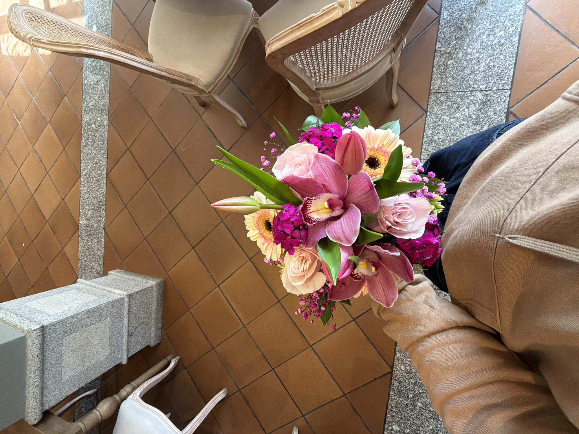 A high-angle shot depicts a person holding a bouquet of flowers in Vila Real, Portugal. The bouquet contains pink orchids, light pink roses, a peach gerbera daisy, and small purple filler flowers. The person is wearing blue jeans and a light brown hooded sweatshirt with the hood down. The background consists of a floor with alternating sections of brown square tiles and gray stone. Two antique-style chairs with light-colored seats and wooden frames are partially visible at the top of the frame. A gray stone block or base is in the lower-left corner of the image, with part of a white chair behind it. The scene appears to be indoors, possibly in a public space or building in Vila Real, Portugal.