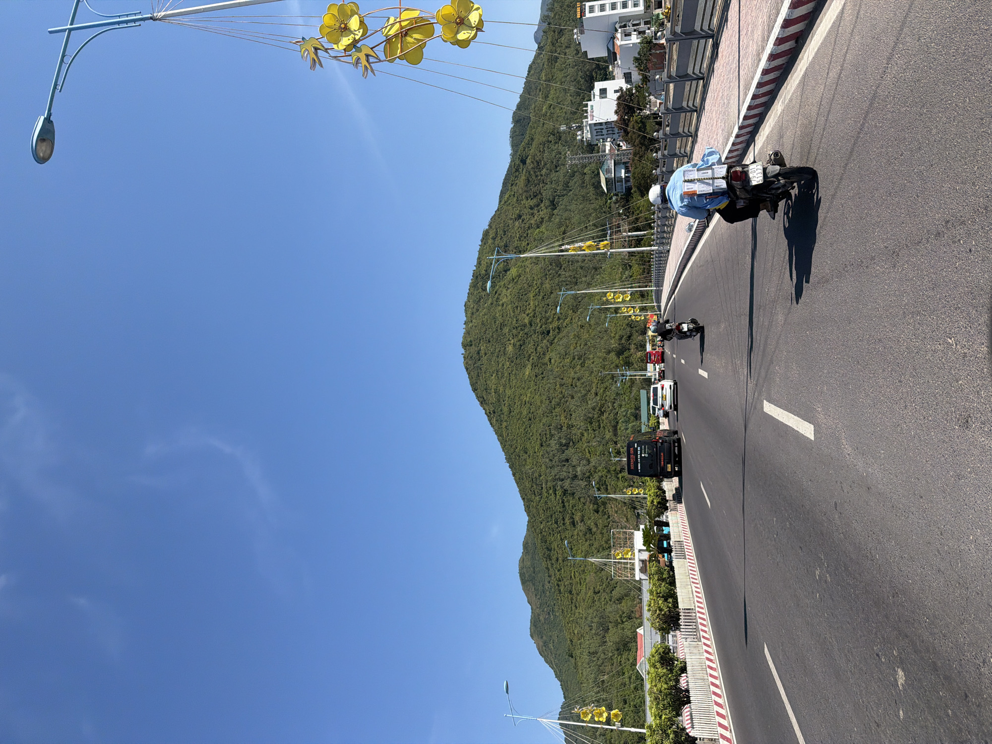 This is a ground-level, eye-level, medium shot of a bridge in Nam Nha Trang Ward, Vietnam, on a sunny day. The bridge is carrying traffic, including a motorcycle in the foreground with a person wearing a light blue shirt and white helmet, carrying packages strapped to the back of the motorcycle. Another motorcycle is visible further ahead on the right side of the road. Cars and a black bus with the text "HERMES SPA" are also on the bridge. The road has clear lane markings. On the left side of the bridge, there is a white fence with red and white striped detailing, and lush green trees. On the right side of the bridge, there is a similar fence and a sidewalk with buildings visible behind it. Tall lampposts with decorative yellow flower-like ornaments line the bridge. In the background, a large, tree-covered mountain dominates the skyline under a clear blue sky.