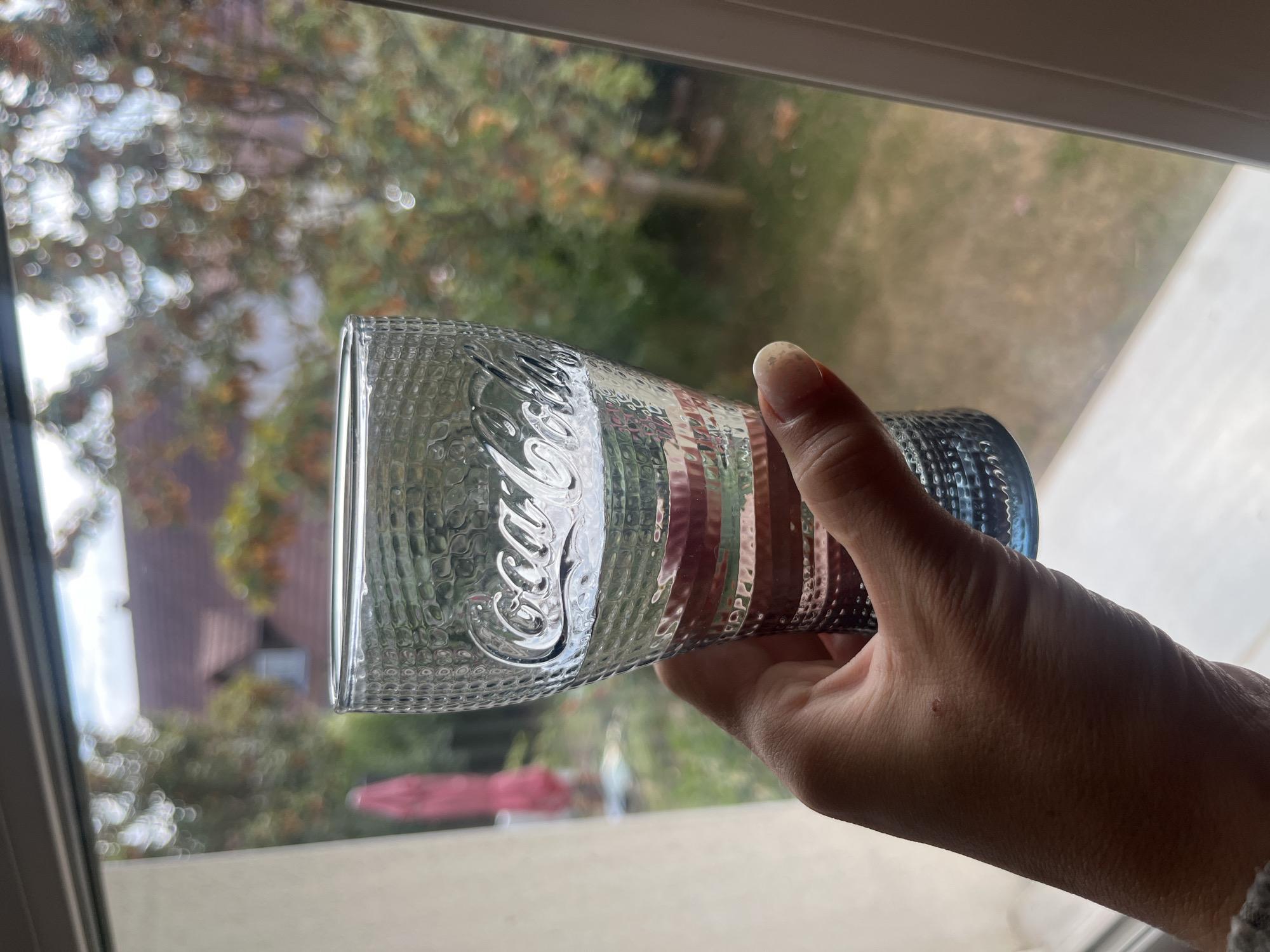 A hand holds a clear, patterned Coca-Cola glass filled with a clear liquid, likely water, in front of a window. The glass has the iconic Coca-Cola logo embossed on it. Outside the window, a blurred background shows trees with autumn foliage, a red umbrella, and hints of buildings. The image appears to be taken in Gertwiller, France.