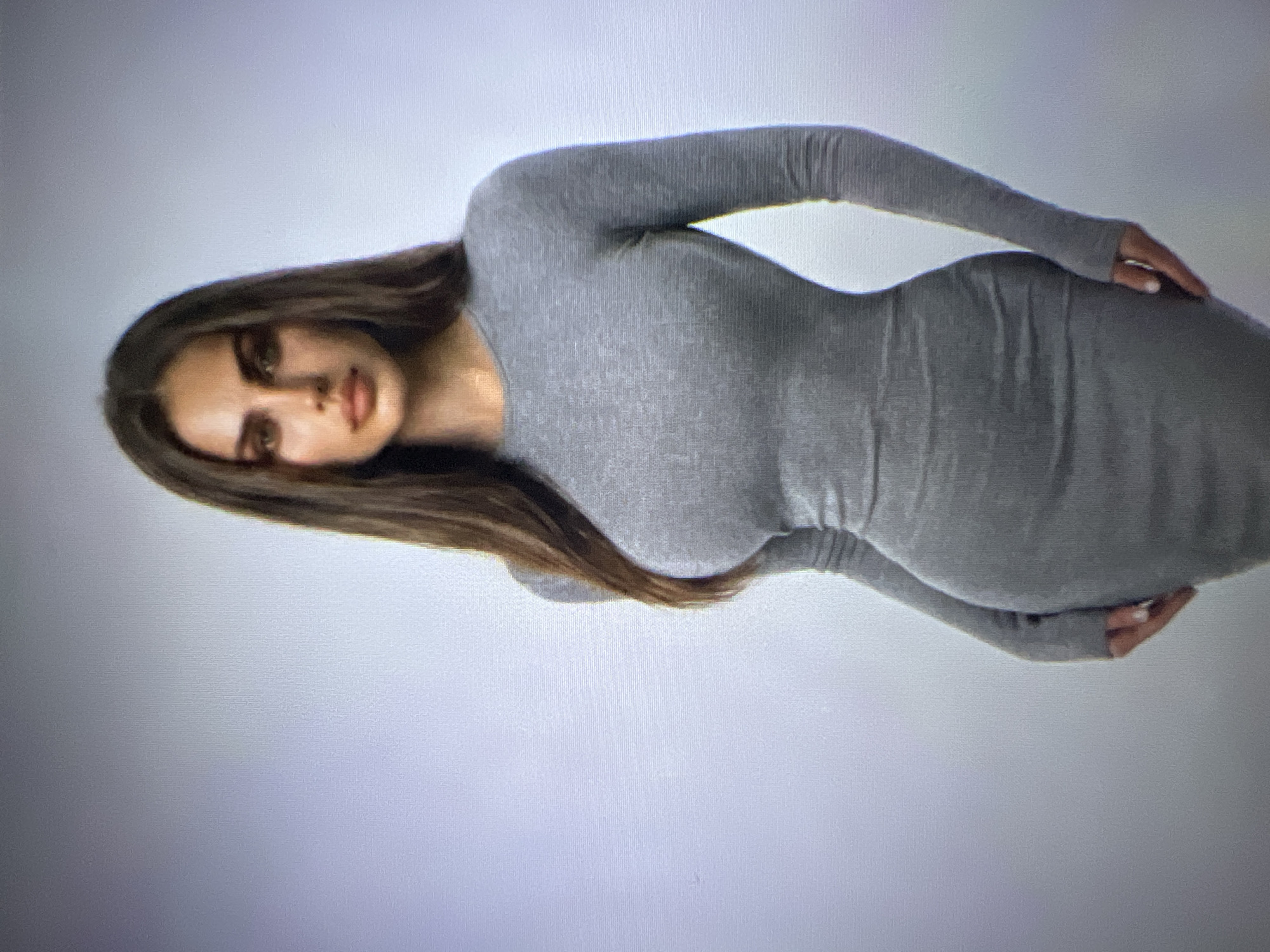 The image shows a full-body studio portrait of a woman with long, dark brown hair. She is wearing a form-fitting, long-sleeved grey dress. The background is a plain, light grey. The lighting is even, and the subject is positioned slightly off-center. There is no indication of any specific location within the city of Pyatigorsk, Russia.
