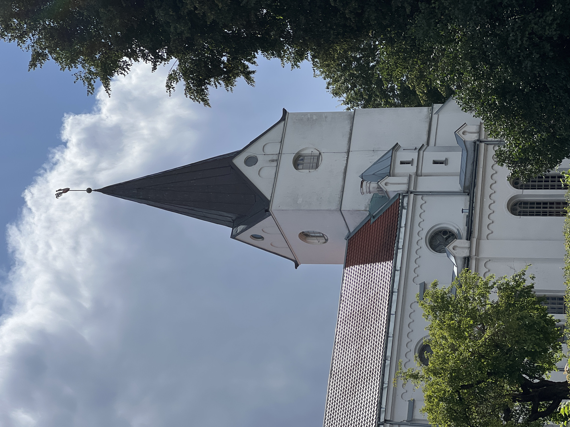 The image shows a close-up of the tower and roofline of the Catholic Church of St. Peter in Saldus, Latvia, on a partly cloudy day. The white church has a dark, pointed steeple topped with a weather vane shaped like a rooster. The roof is covered with red tiles. Lush green trees flank the right side of the church. The sky is a mix of blue and dramatic white clouds.