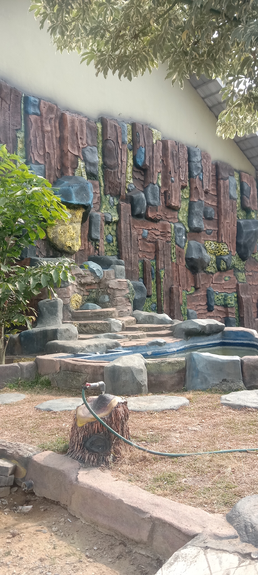 The image shows an artificial rock formation with a waterfall and a small pond. The rock formation is decorated with various colors and textures, mimicking natural rock. There is a tree with green leaves on the left side of the image. In the foreground, there is a tree stump with a tap and a hose. The ground is covered with dry grass and some stones. The scene appears to be located in a park or a zoo. There are no people visible in the image. The overall impression is that of a man-made attraction designed to resemble a natural landscape. The location in the city of Sumber Agung Mataram, Indonesia, is not explicitly visible in the image, but this is a general description of the scene.
