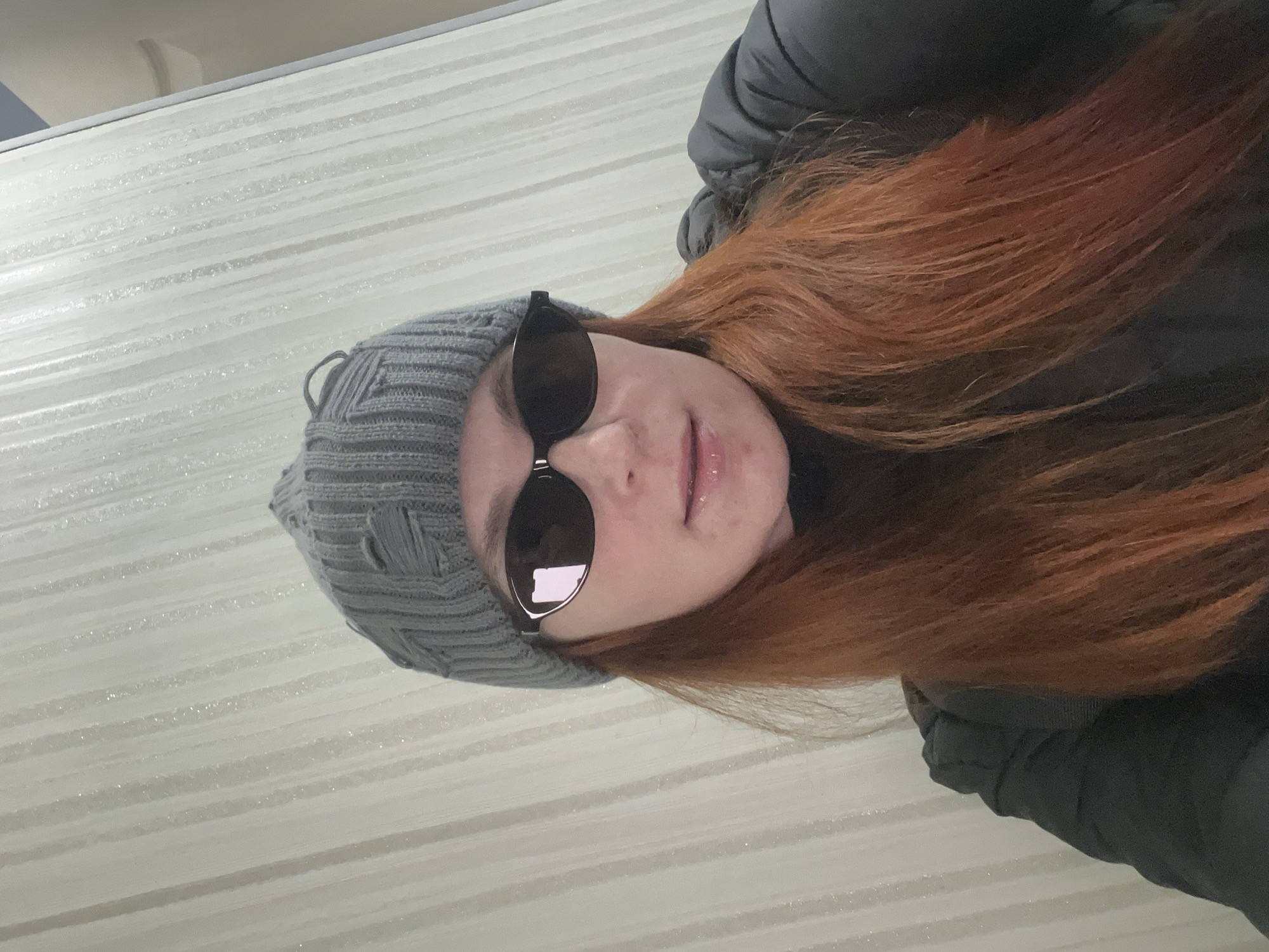 The image features a person wearing a grey knitted beanie and sunglasses. The person has red hair and is wearing a dark-colored jacket. The background appears to be a textured, striped wall. There is no location information provided in the image.