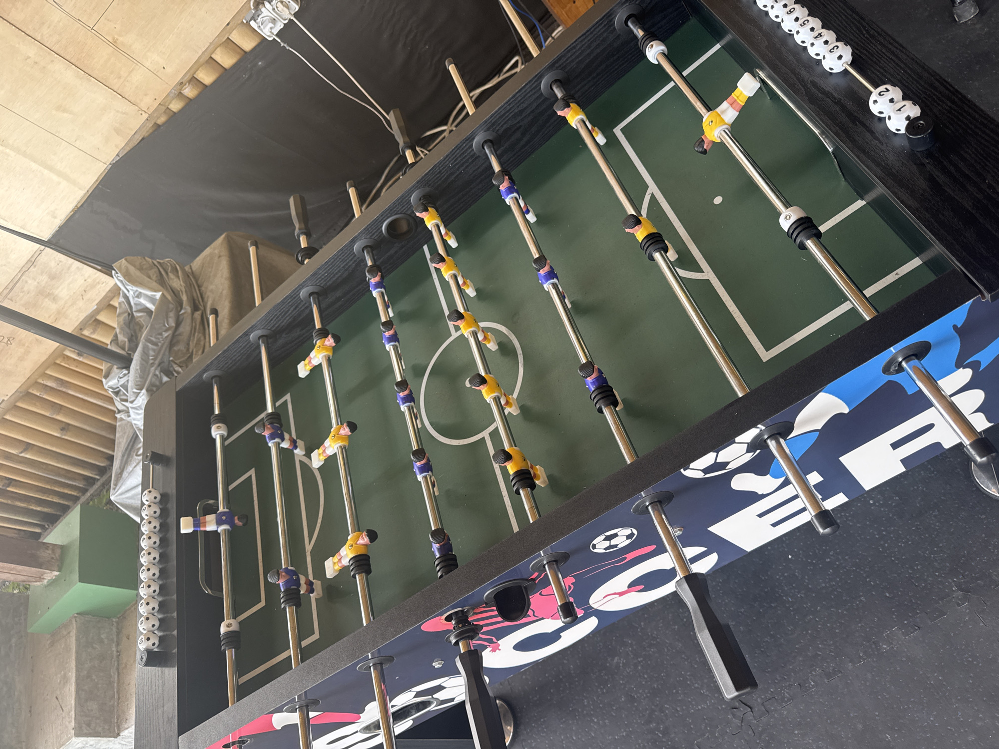The image shows a foosball table in an indoor setting. The table features a green playing surface with white lines mimicking a soccer field. Silver metal rods extend across the table, with plastic figures in yellow and blue attached to them, representing players. Black handles are visible on the near side, used for controlling the figures. The table has a decorative blue and white design around its edges, featuring soccer ball patterns. White and black score counters are mounted on the ends of the table. The environment appears to be a semi-covered area, with a glimpse of what may be outdoor foliage visible in the background. The location, Nusa Penida, Indonesia, is not apparent within the visual contents of the image.