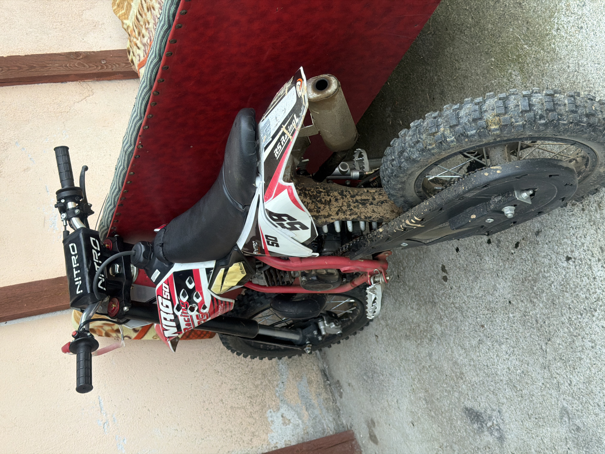This is a close-up, slightly tilted, outdoor, eye-level shot of a dirt bike. The dirt bike is red and white with black accents. It is leaning against a textured red surface, possibly a seating area or a tarp. The ground beneath the bike appears to be concrete. The bike is covered in some dirt, suggesting recent use.
The visible text on the bike includes "NITRO" printed vertically on a black section near the handlebars, and "NRG 50 Racing" on a graphic decal. There is also a "50" graphic on the side.
The background shows a textured wall, likely part of a building, with a wooden beam visible. The lighting suggests it is daytime. There are no people visible in the image. The scene is static, with the dirt bike appearing to be parked or at rest. The overall impression is one of a used recreational vehicle in a casual outdoor setting.