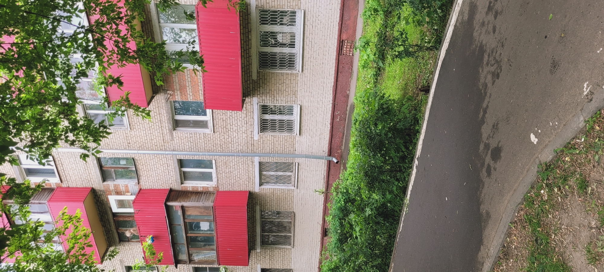 The image shows a multi-story brick apartment building in an urban environment, presumably in the city of Unknown. The building features numerous windows, some with metal security bars. Many balconies have red corrugated metal roofs. The building is adjacent to a sidewalk, with green foliage growing between the building and the walkway. A paved road or path is visible to the right of the sidewalk. The image appears to have been taken during daylight hours. No people are visible.