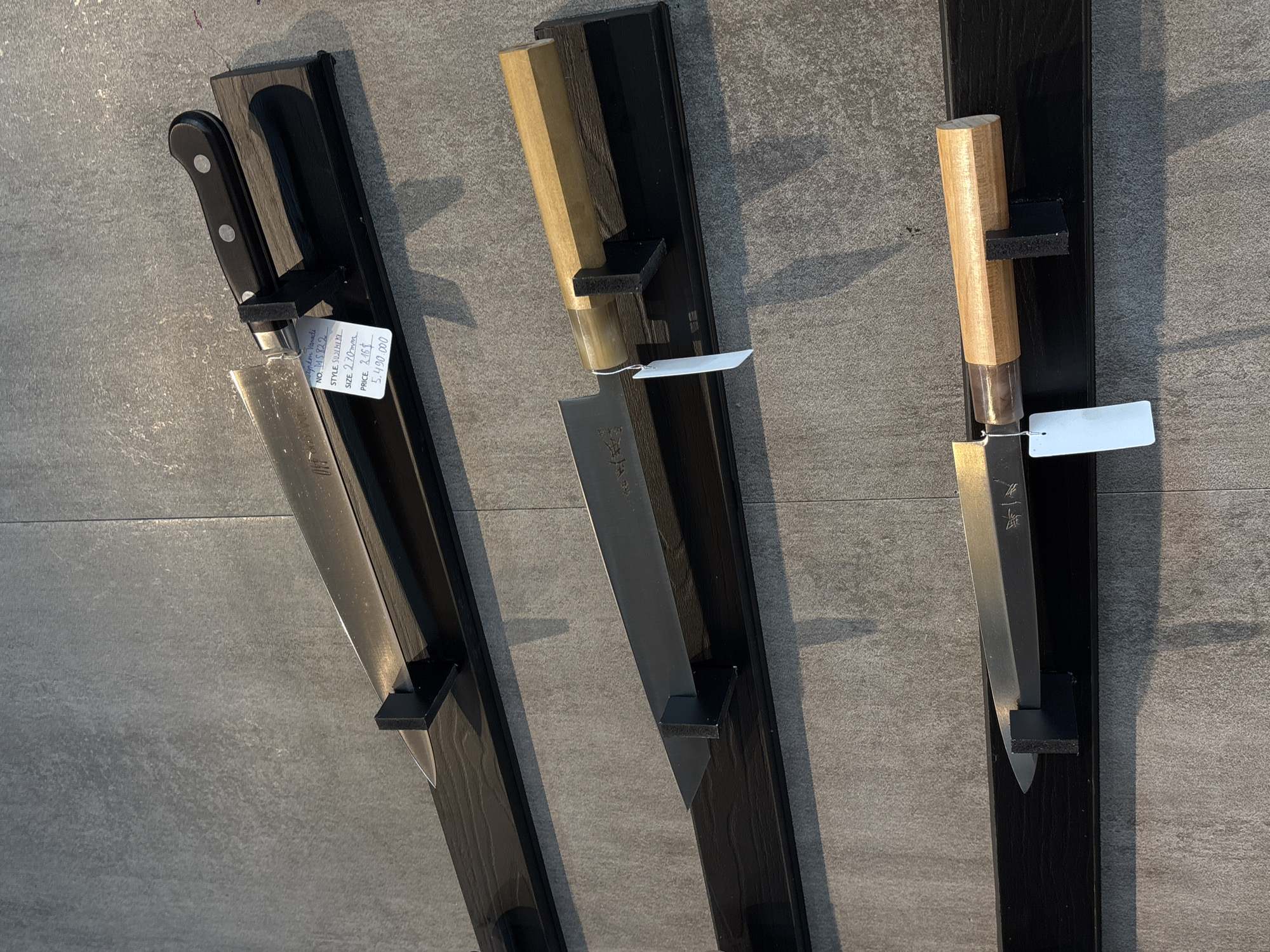 Three knives are displayed vertically on individual dark mounts against a textured grey wall, likely in a retail setting in Đà Nẵng, Vietnam. The leftmost knife features a black handle with rivets and a wide, possibly stainless steel blade, secured by a clear blade protector. A white price tag hangs from its bolster. The middle and rightmost knives have light wooden, octagonal-shaped handles and narrower blades, also with clear protectors and small white tags. The blades of the middle and right knives appear to have etched or stamped Japanese characters or symbols near the bolster. Shadows are cast on the wall, indicating an indoor lighting source.
Visible text on the price tag of the leftmost knife reads:
"Agpton Vanadi"
"NO. MS822"
"STYLE. SOLAIKI"
"SIZE. 2.70mm" (likely referring to blade thickness, though "270mm" for length is common for knives)
"PRICE. 216$"
"5.490.000" (Vietnamese Dong, confirming the location context).