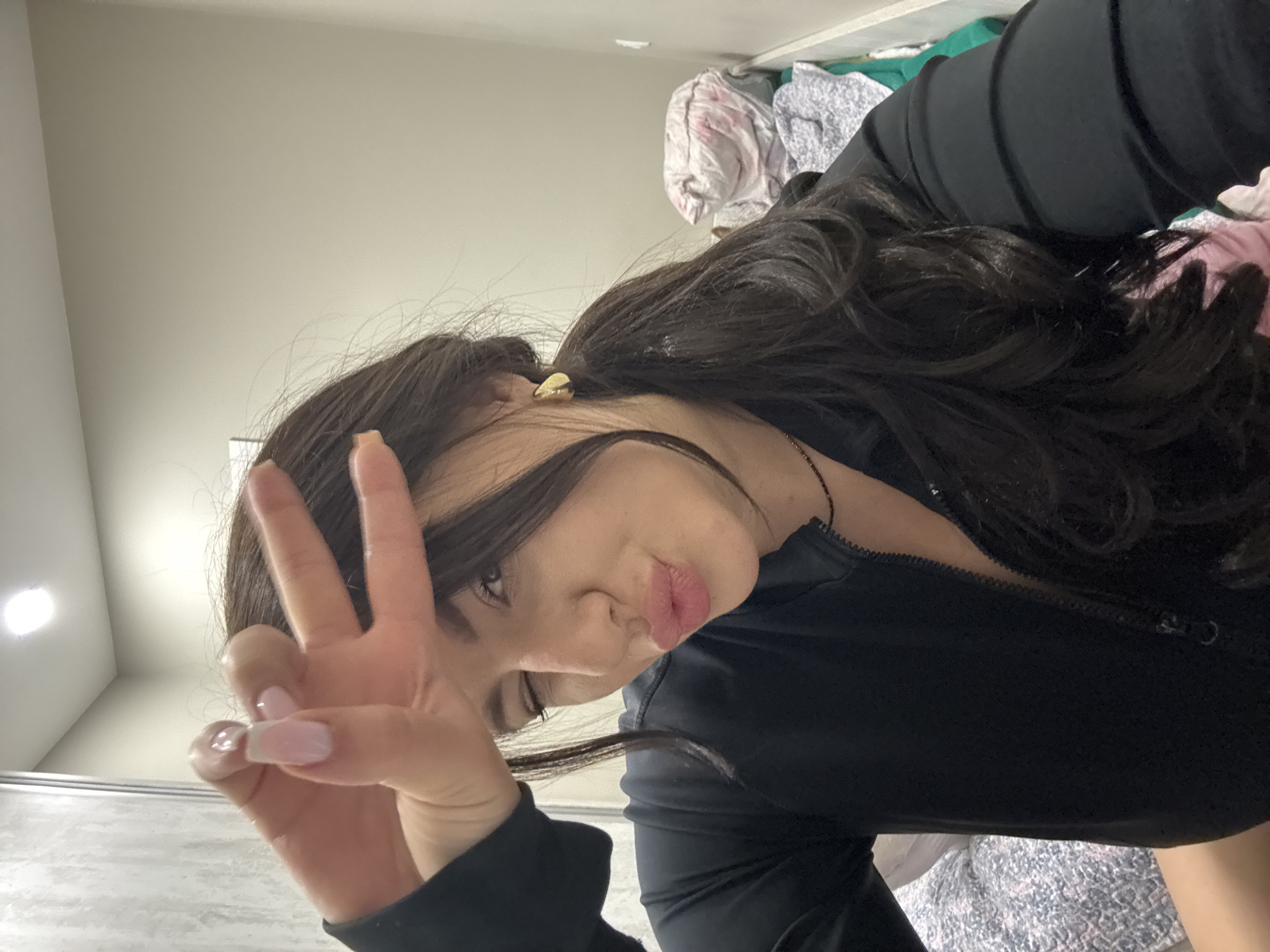 A young woman with long, dark, wavy hair is captured in a close-up, rotated image, appearing to be a selfie taken indoors. She is seen from the chest up, with her head tilted to the right and her gaze directed towards the camera. Her left hand is raised, making a peace sign (V-sign) with her index and middle fingers, revealing nails with light pink French tips. Her lips are pursed into a "duck face" or kissy face, conveying a playful or confident expression.
She is wearing a black, long-sleeved top with a visible zipper down the front. A small, golden-colored earring is visible in her right ear, partially obscured by her hair.
The background suggests an indoor setting. Behind her, a plain, light-colored (beige or off-white) wall is visible, with a light fixture on the ceiling towards the upper left. To the right, behind her shoulder, there is a cluttered area with various articles of clothing, including items in pink, white, green, and grey patterns, possibly piled or folded. The lower part of the background on the left shows a darker, possibly metallic or tiled surface. There is no visible text in the image.