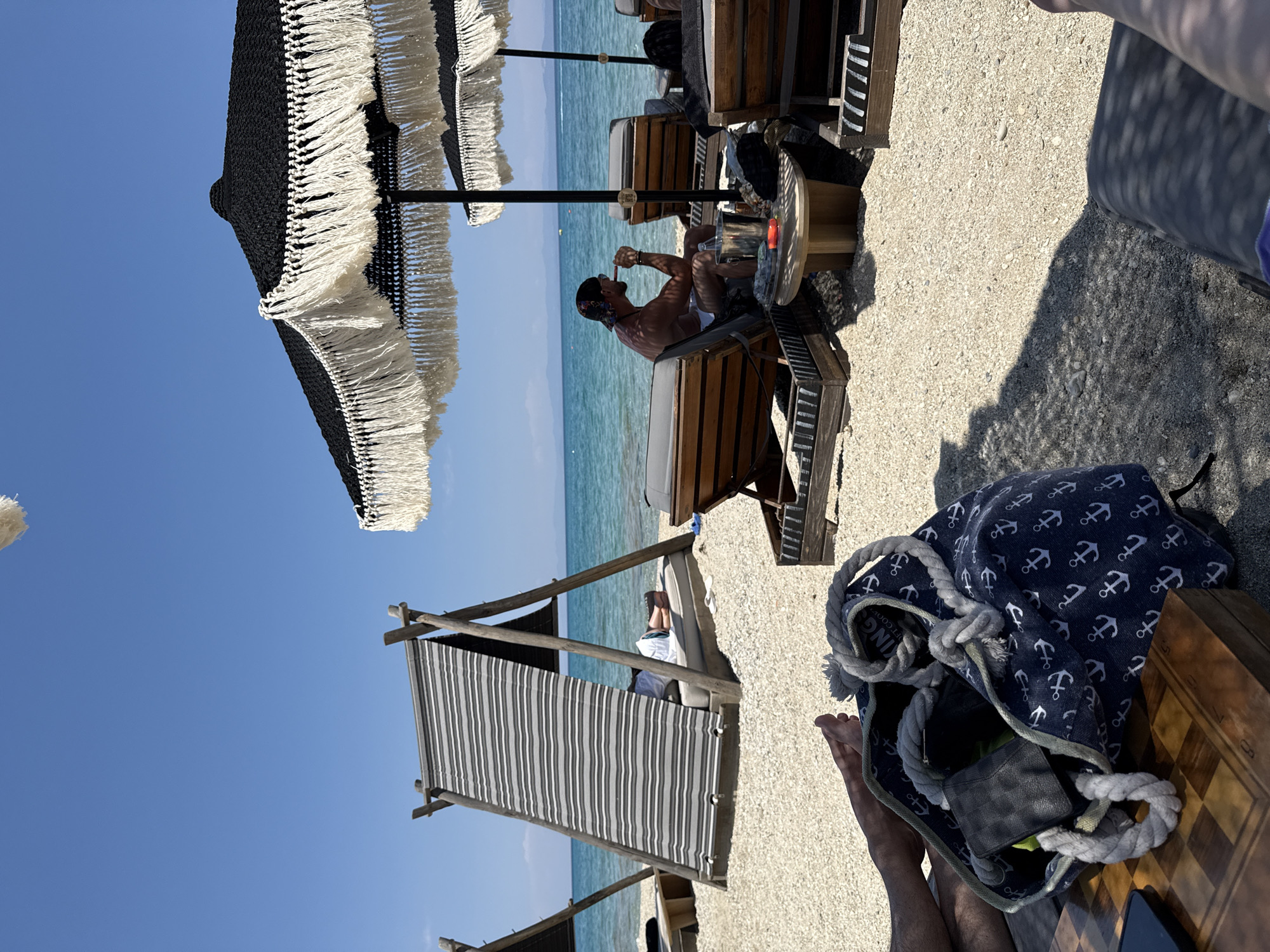The image depicts a sunny day at a beach, likely in Pallini Municipal Unit, Greece. The foreground shows a person's legs and feet, along with a dark blue tote bag adorned with white anchor patterns. The bag contains a wallet and possibly other items. A wooden chessboard is also visible next to the bag.
In the midground, a man is relaxing on a sun lounger under a black and white fringed umbrella. He is holding something to his mouth, perhaps a drink or food. The sun lounger is made of wood and has grey cushions. Next to him, a small wooden table holds a glass and a bottle. The background features a calm, turquoise sea and a clear blue sky. Another striped cabana structure is visible further down the beach. The overall atmosphere suggests a leisurely and relaxing day at the beach. The lighting indicates it is daytime, likely late morning or early afternoon, given the strong sunlight and clear skies.