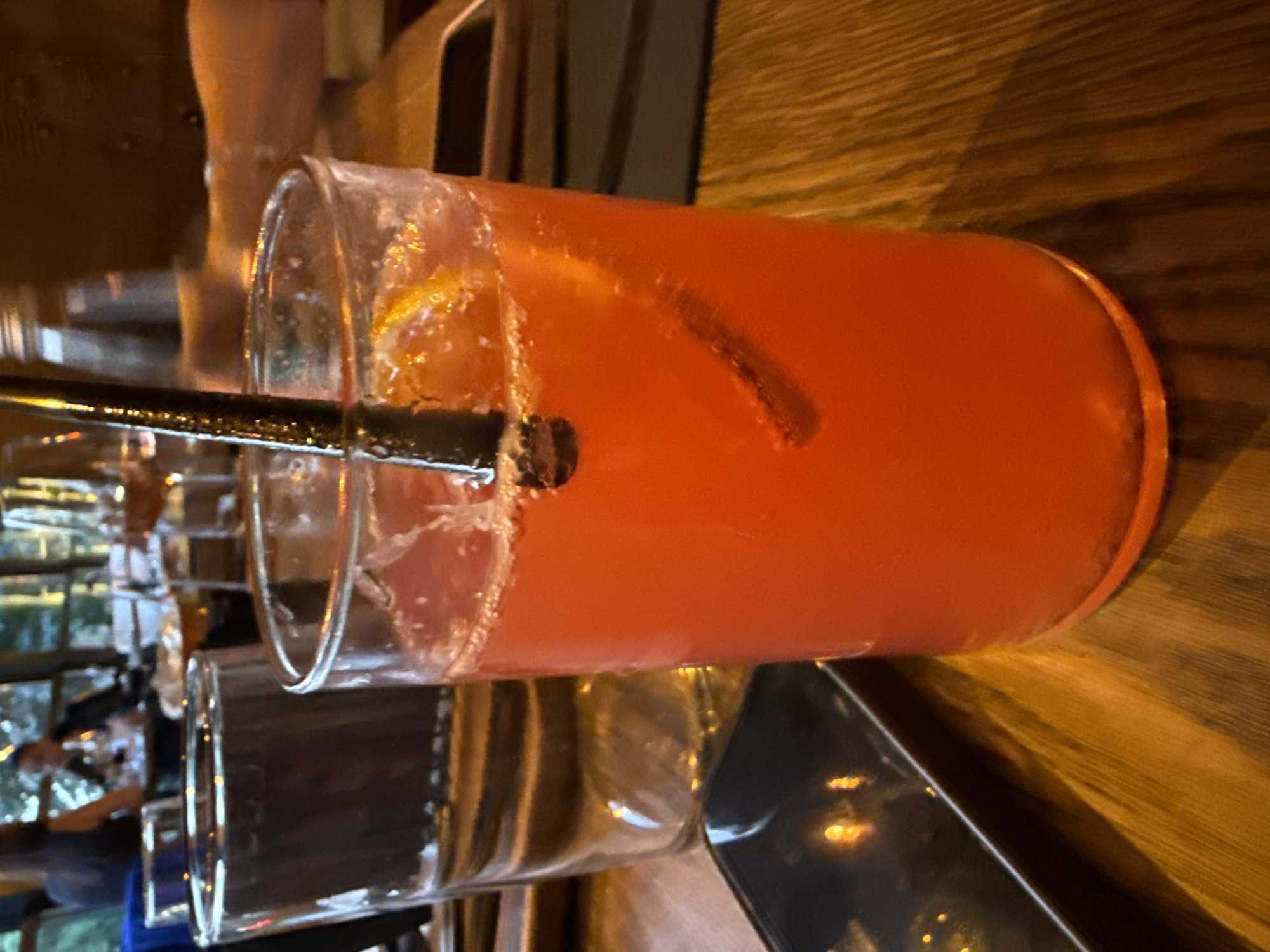 A close-up, eye-level shot captures a vibrant orange-red cocktail in a clear glass, adorned with a straw and a slice of fruit, set against a wooden table. The drink is a focal point, its effervescence and bright hue suggesting a refreshing beverage. In the background, out of focus, are other patrons and glasses, hinting at a lively restaurant or bar setting. The lighting is warm and ambient, casting a soft glow on the scene. The overall impression is one of a relaxed evening, possibly in a metropolitan area. This particular setting is identified as being in Sofia, Bulgaria, suggesting a possible dining or social gathering experience within the city.