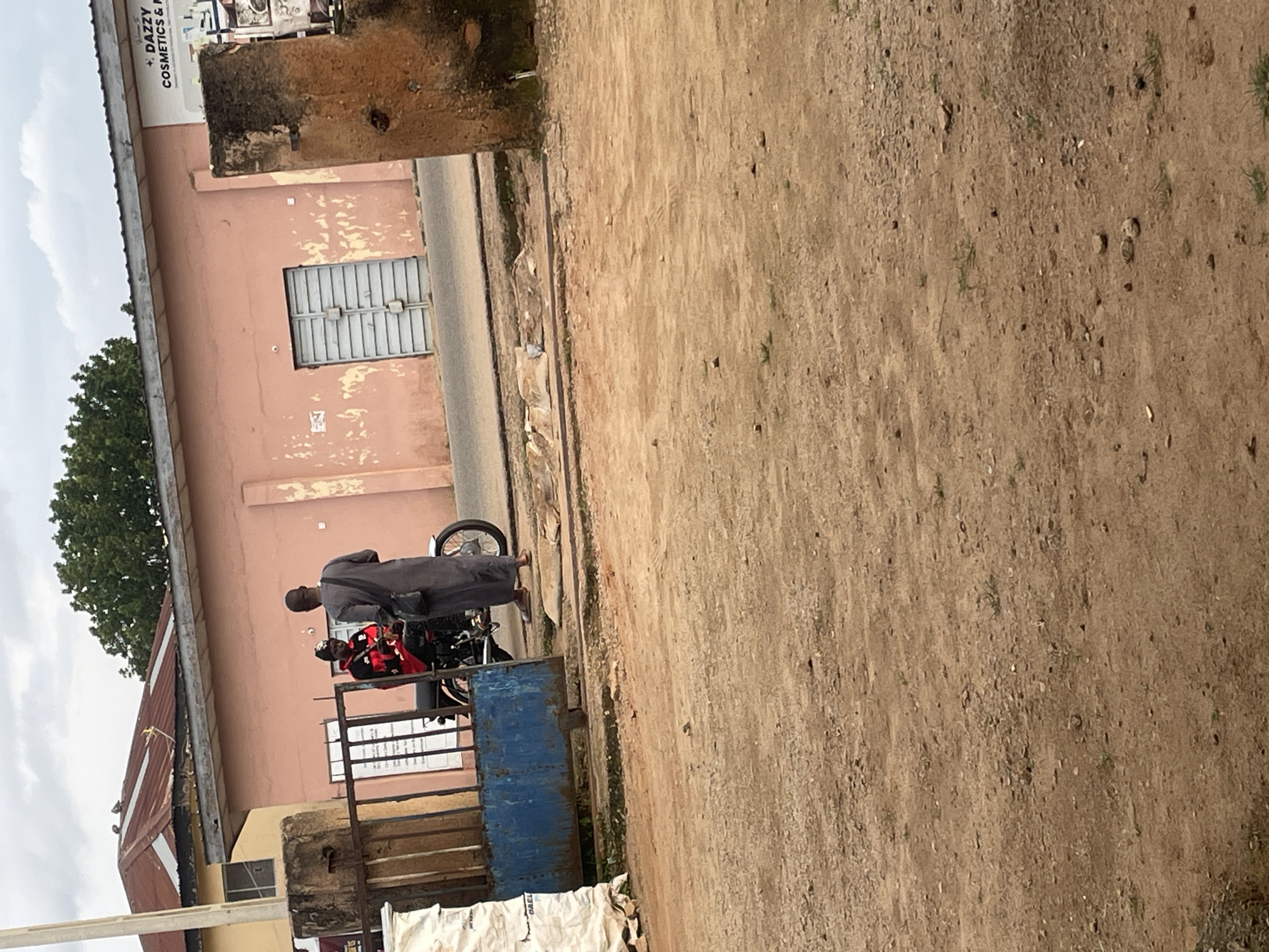 A male individual wearing a dark gray robe and a black shoulder bag stands on a dirt road beside a blue metal gate. He is facing a motorcycle that has a male rider wearing a red jacket with black accents and a black beanie. The motorcycle is positioned partially inside the gate, and the rider appears to be holding a phone. Behind the gate, a light pink building with a corrugated metal roof is visible. The building has several windows and a large metal door. A sign is mounted on the wall of the building that reads "+ DAZZY COSMETICS & P". The surrounding environment is characterized by a dusty, dirt ground, sparse vegetation, and an overcast sky. The scene appears to be in a commercial or semi-industrial area, possibly within the city of Jos, Nigeria, given the architectural style and common modes of transportation.