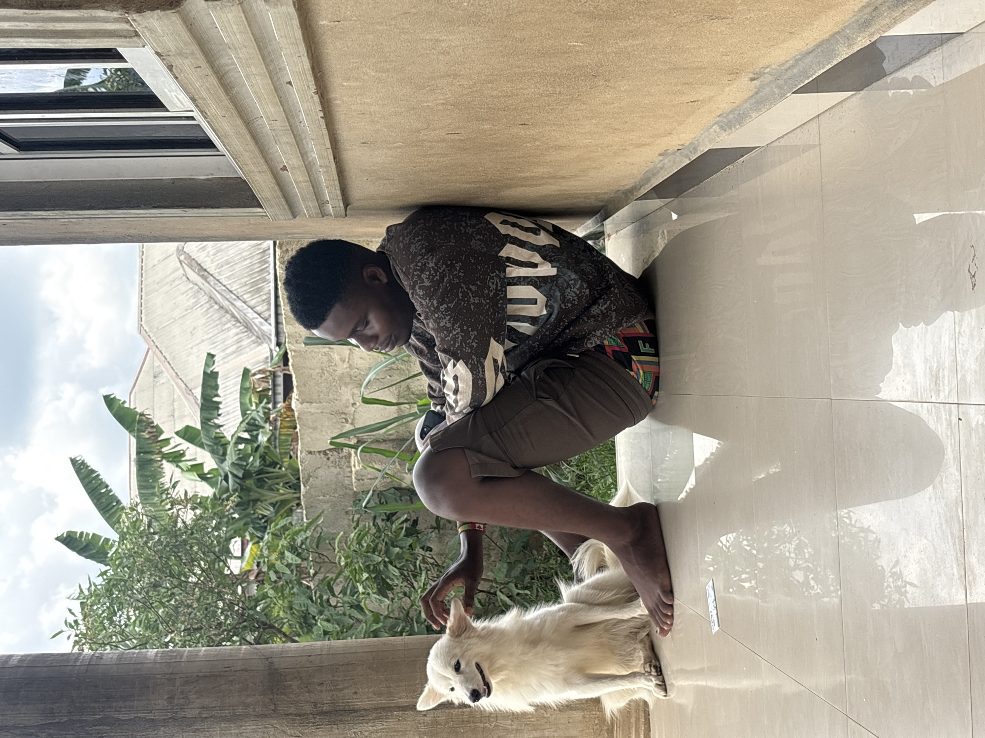 Outside a building in Ikot Ekpene, Nigeria, a young man squats on a tiled surface, facing and petting a medium-sized, white dog. The man wears a dark patterned sweatshirt and dark shorts, and has a colorful bracelet on his left wrist. The dog lies on the ground, looking up at the man. In the background are plants, part of a building, and the sky. The light source is natural, casting shadows across the scene.