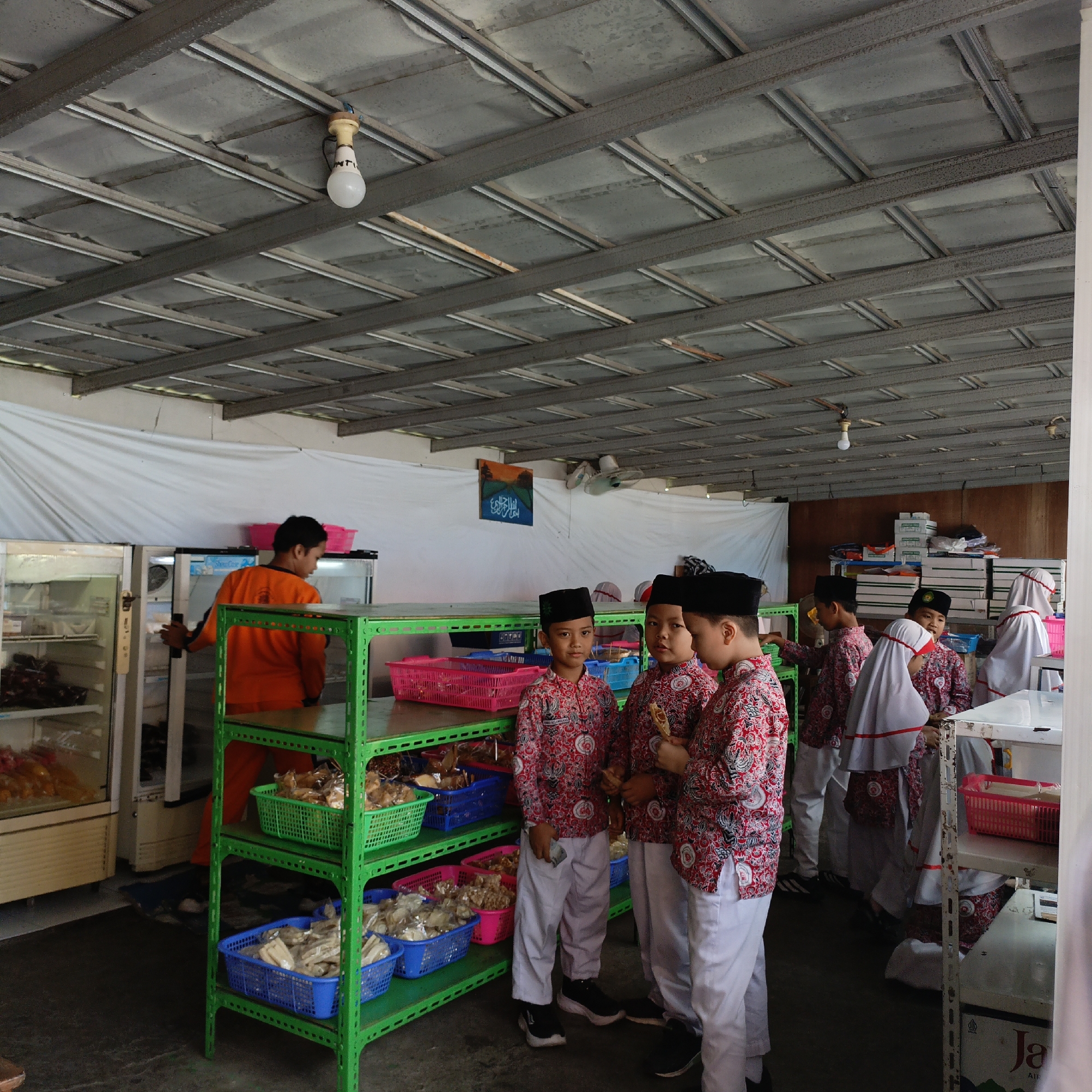 This image depicts a group of young boys, likely students, dressed in traditional attire including white peci caps and patterned shirts, standing in what appears to be a covered outdoor market or food stall area in Salatiga, Indonesia. They are gathered near a bright green metal shelf unit stocked with various food items in baskets. To the left, there are commercial refrigerators, one of which is open, revealing more products. A person in an orange uniform is visible near the refrigerators. The ceiling is constructed of metal beams and panels, with a lightbulb hanging from the center. The overall environment suggests a casual, possibly commercial, setting.