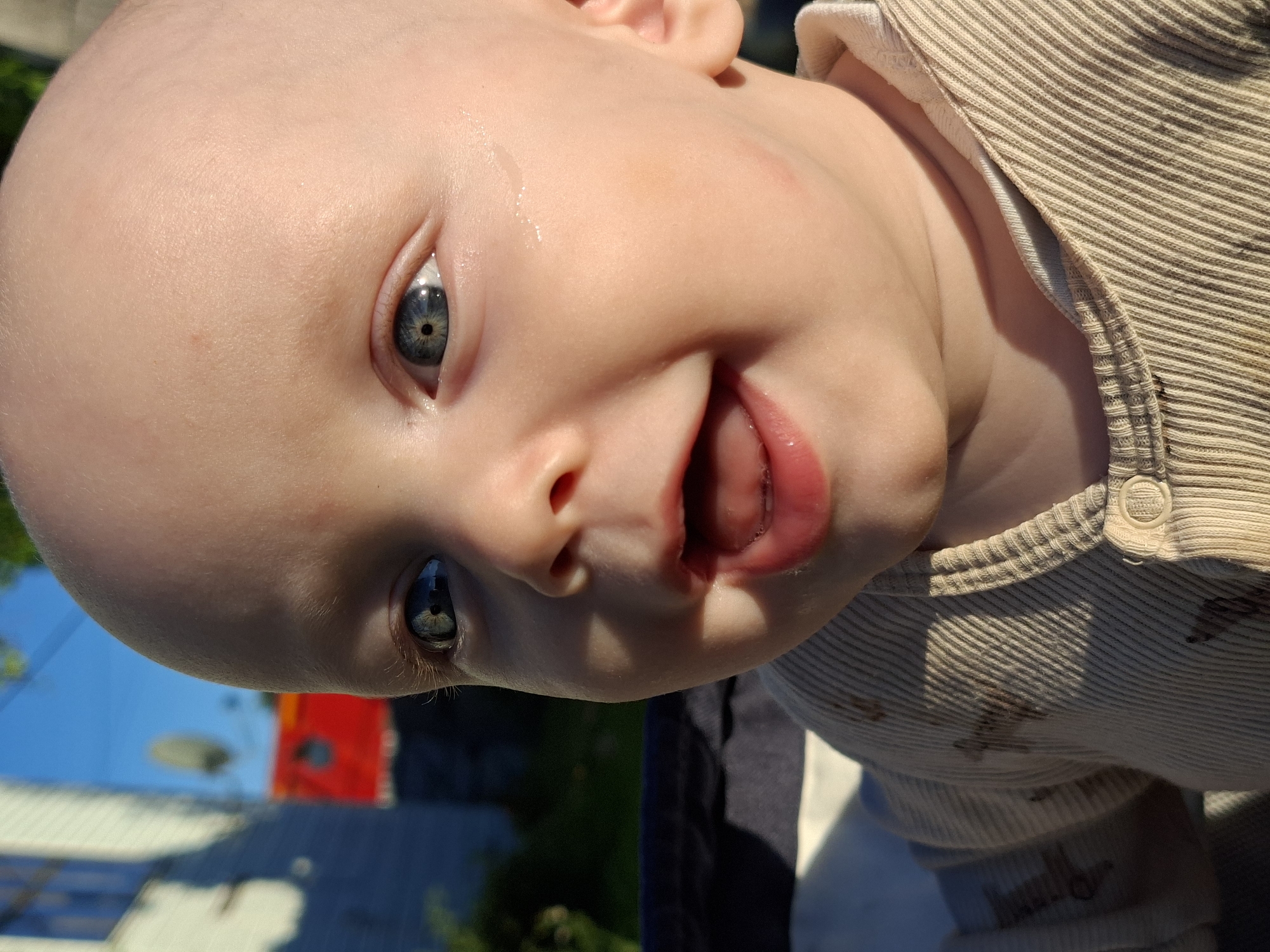 An extreme close-up shot captures a baby's face from the side, bathed in golden sunlight. The baby, with a bald head and wide, blue eyes, is looking to the left with a joyful, open-mouthed expression. A small bead of sweat glistens on the baby's cheek. The baby is wearing a light beige, ribbed shirt with a collar. In the background, out of focus, a light-colored building with a dark window and a red structure with a circular opening are visible under a bright blue sky. The setting appears to be outdoors.