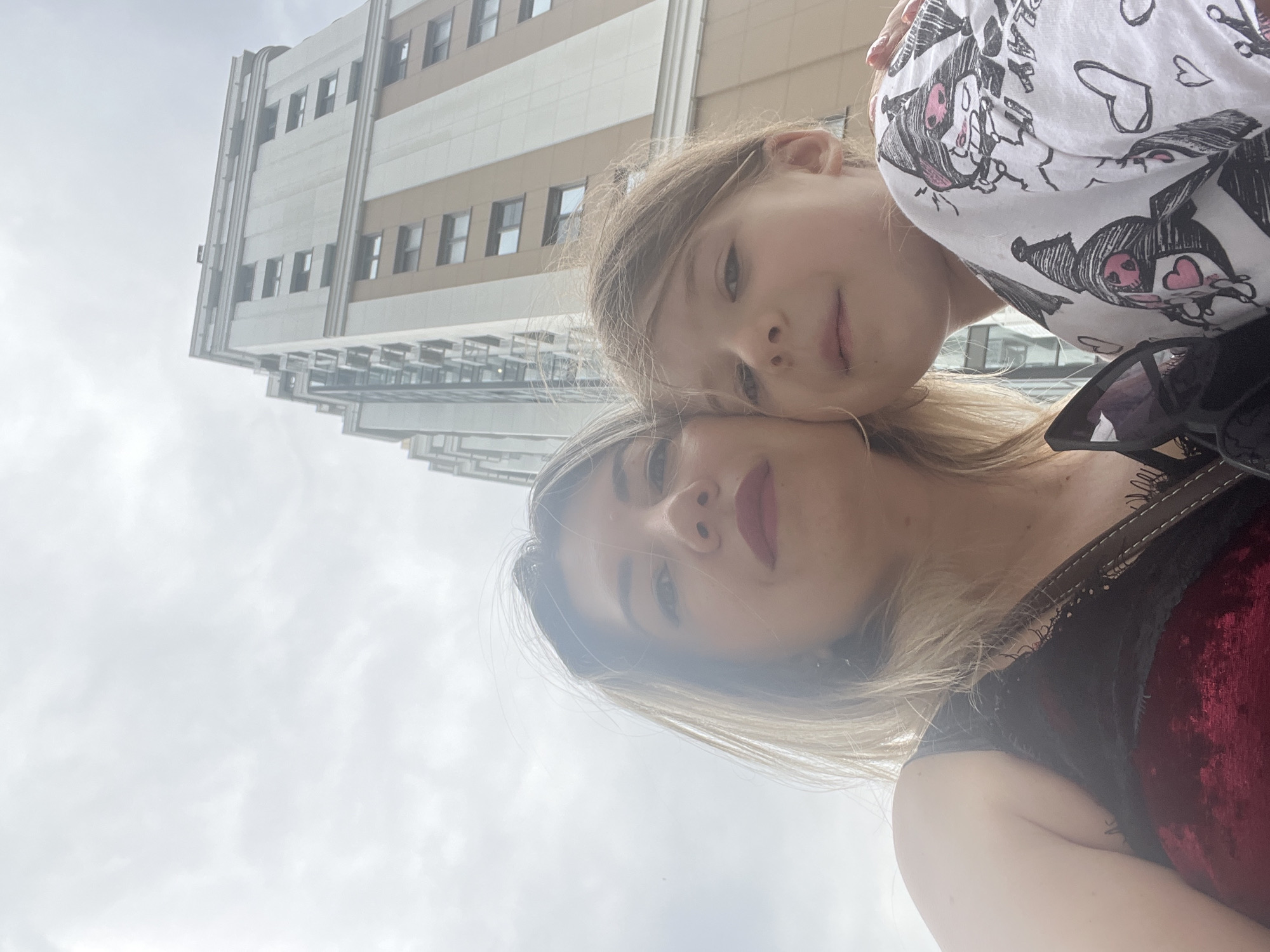A low-angle, medium shot captures a woman and a young girl in front of a tall, modern apartment building against a cloudy sky. The woman is on the left, her face partially obscured by a lens flare, with blonde hair and dark lipstick. She is wearing a dark red velvet top with black lace trim. The girl is on the right, looking forward, with blonde hair and wearing a white t-shirt with black cartoon drawings. A pair of sunglasses rests on the woman's shoulder. The apartment building is light beige and white, with multiple rows of windows. The setting is the city of Sochi, Russia, indicated by the architectural style of the building and the general atmosphere.