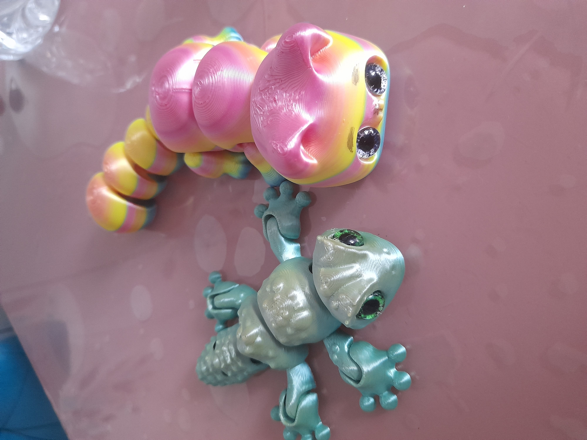 The image depicts two 3D-printed articulated toys resting on a maroon-colored surface. At the top is a multi-colored caterpillar-like toy with a yellow, pink, and blue gradient. It has a rounded head with two prominent black eyes. Below, a light blue-green articulated lizard toy is positioned with its limbs spread out. The lizard also has inset eyes. The scene appears to be indoors, with some blurred details possibly indicating reflections or other objects in the background. The city and location are unknown.