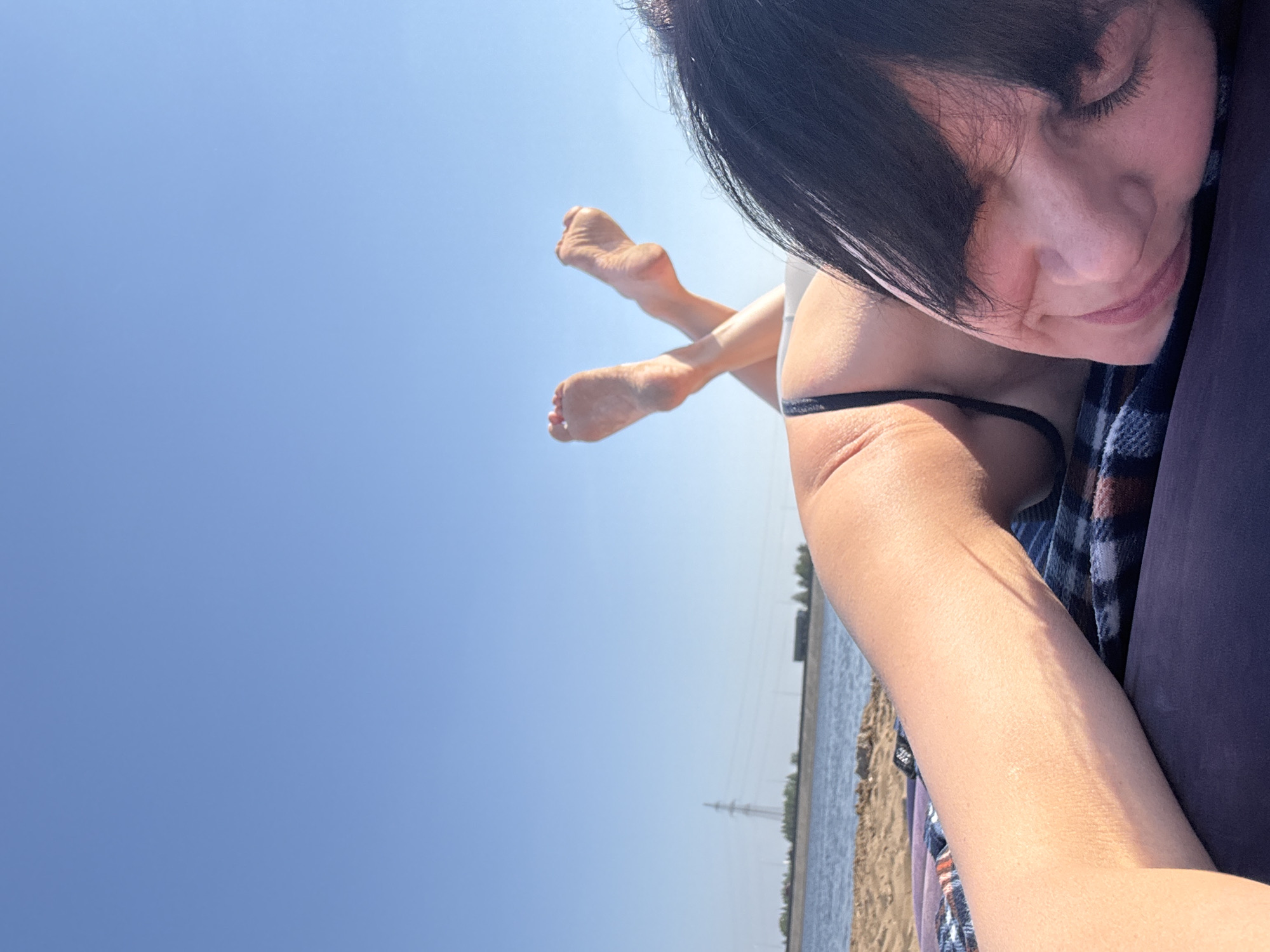 A woman is lying on a blanket by the water, possibly on a beach, in what appears to be Chaikovsky, Russia. Her face, with closed eyes and black hair, is in the upper right corner of the frame. Her legs, crossed at the ankles, are pointing upward toward the bright blue sky. A body of water is visible with power lines and towers in the distance. The woman appears to be wearing a bathing suit. She is lying on a blanket.