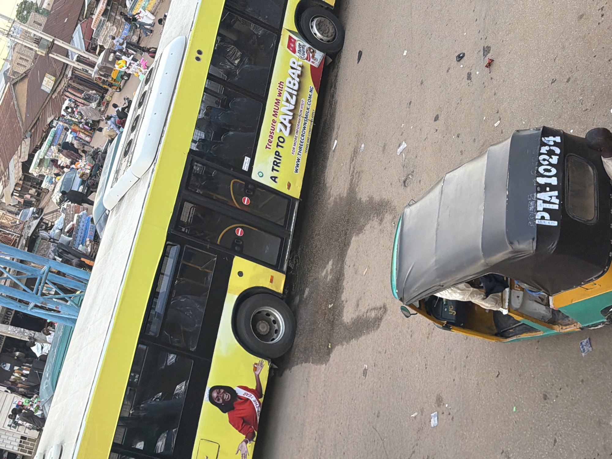 This is a ground-level, slightly elevated, full-shot photograph taken in Bukuru, Nigeria, focusing on a yellow public bus and a three-wheeled vehicle known as a "keke" in the foreground. The bus is parked on an asphalt road, partially obstructing the view of a busy marketplace situated behind it. The marketplace appears to be a vibrant commercial hub with numerous stalls displaying goods, including textiles and mattresses. The bus itself is decorated with advertisements, most notably one featuring a woman in a red dress and a sash that reads "MISS YEAR". Another advertisement on the side of the bus promotes a trip to Zanzibar and a product called "Three Crowns Milk". The keke in the foreground is green and yellow, with the license plate PTA-10234 clearly visible. The driver of the keke is wearing a white traditional garment. Several people can be seen in the background, engaged in various activities such as walking, shopping, and operating motorcycles. The overall scene depicts a typical day in a bustling Nigerian city, with a strong emphasis on transportation and commerce.