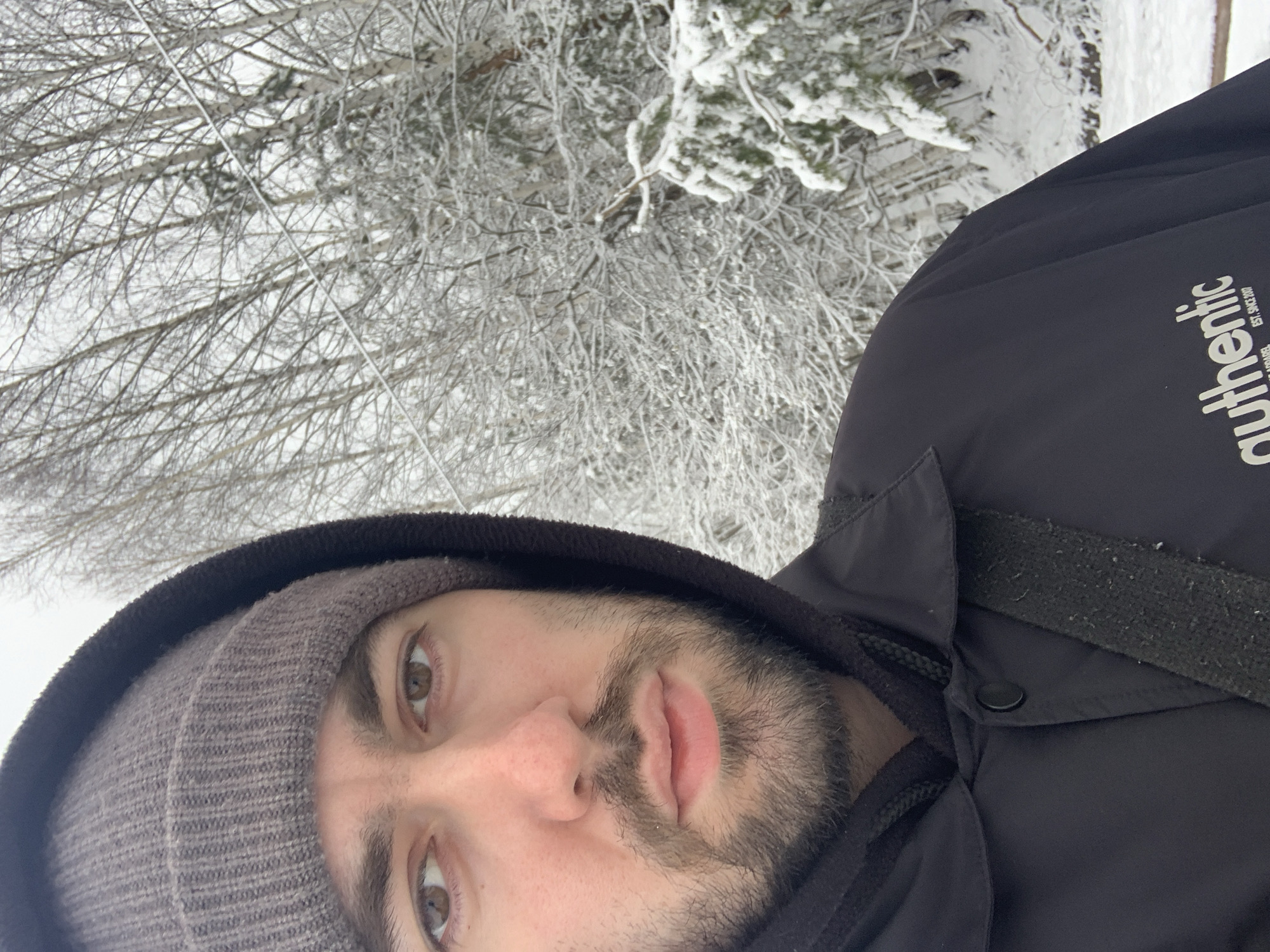 A close-up selfie photograph captures a bearded man with a tan beanie and dark hood, wearing a dark jacket with "authentic" text visible on the shoulder. He has brown eyes and light skin. The background shows a snowy forest scene, potentially taken during winter. It is not possible to confirm the location as the city of Chaikovsky, Russia, from the visual information alone.