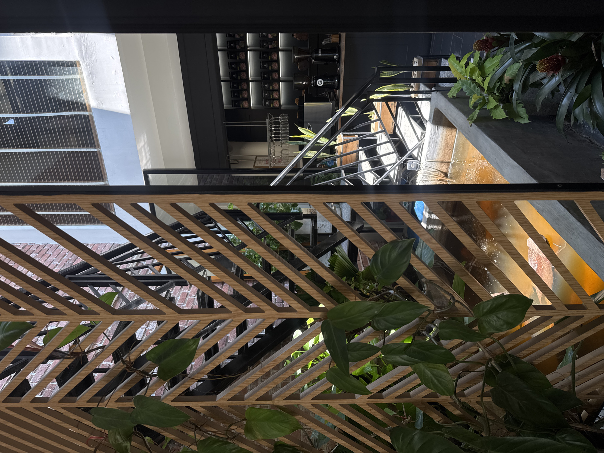 The image shows an interior view through a lattice-style wooden screen, with greenery partially obscuring the screen. Behind the screen, there is a view of a building with a modern interior design. This interior includes a metal staircase, a water feature or fountain, and shelves containing coffee equipment or supplies. The building itself appears to be of brick construction. The setting is Unknown, Unknown.