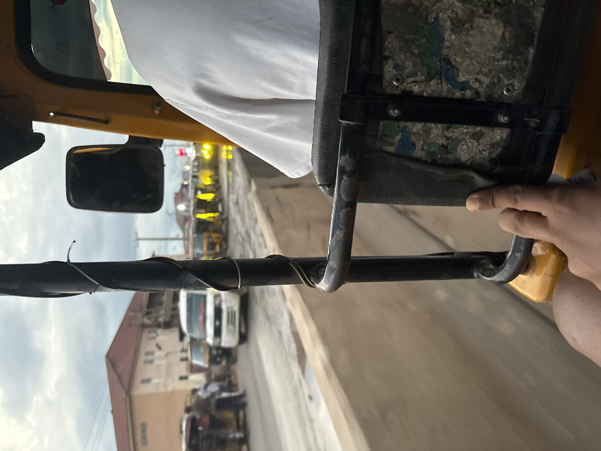 The image captures a view from inside a yellow auto-rickshaw in Onitsha, Nigeria. The foreground shows the right hand of the passenger resting on the edge of the auto-rickshaw's seat, which has a dark textured surface with a patterned inlay. A black metal bar, part of the auto-rickshaw's structure, is prominently visible. Beyond the auto-rickshaw, a street scene unfolds under an overcast sky. Several other auto-rickshaws and a white van are present on the road, suggesting traffic congestion. In the background, a two-story building with a reddish-brown roof stands. The environment appears to be an urban setting, possibly a market area or a busy road in Onitsha, a major commercial hub in Nigeria.