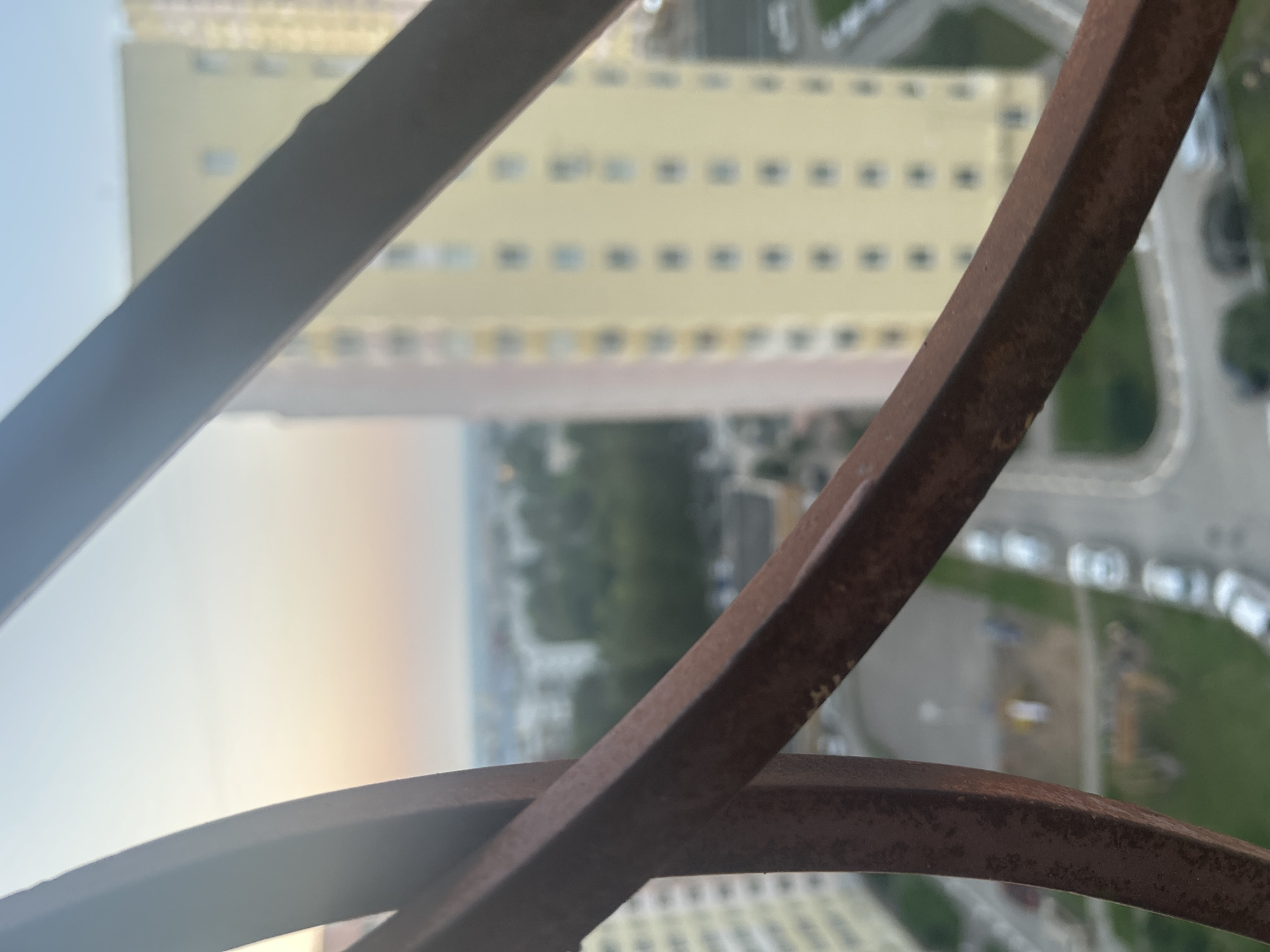 The image, taken from a high vantage point, shows a blurred urban landscape with tall, light-yellow apartment buildings and greenery. In the foreground, rusty metal bars obscure part of the view. The sky in the background displays a gradient of soft colors, suggesting either sunrise or sunset. The scene appears to be a residential area within a city, possibly in Perm, Russia, given the architectural style of the buildings and the general appearance of the urban environment. There are no identifiable people or specific actions depicted.