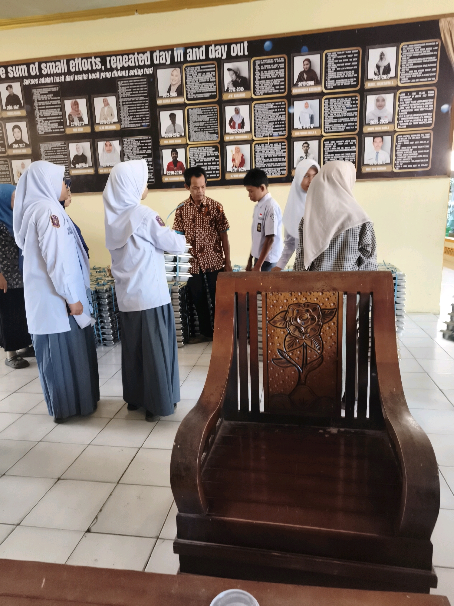 The photograph captures an indoor scene, likely a classroom or meeting room, with a large bulletin board displaying portraits and names. The bulletin board features text at the top reading "the sum of small efforts, repeated day in and day out" followed by "Sukses adalah hasil dari usaha kecil yang diulang setiap hari". Below this, numerous framed photographs of individuals, each with accompanying text, are arranged in a grid.
In the foreground, a dark wooden chair with intricate carvings of a flower and leaves is prominent. To the left of the chair, four young women, presumably students, are standing. They are all wearing similar light-colored long-sleeved shirts and long grey skirts. Three of the women are wearing headscarves. One of the women on the far left is wearing a black patterned garment and a dark headscarf.
Standing behind the students, in the middle of the frame, is a man wearing a patterned short-sleeved shirt and dark pants. He appears to be addressing the students. To the right of the man and behind the students, stacks of rectangular metal containers are visible. The room has tiled flooring. The overall impression is of an educational setting where achievements are recognized, possibly at a school in Makassar, Indonesia, given the Indonesian text on the bulletin board.