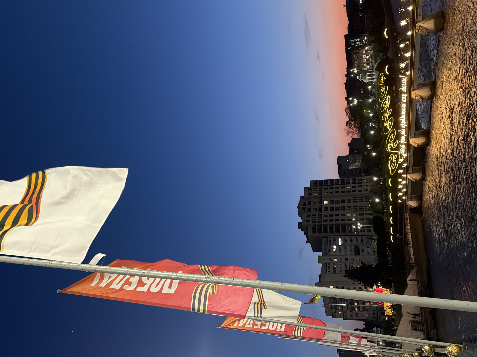 The photo, taken in the city of Chermen, Russia, shows a row of celebratory flags fluttering in the foreground against a backdrop of buildings illuminated with evening lights. The flags feature the St. George ribbon, a symbol associated with Victory Day, and the Russian word "ПОБЕДА" (POBEDA), meaning "Victory".
The sky transitions from a deep blue to a soft pink-orange hue near the horizon. A bridge, adorned with decorative lights and script in Russian that translates to "This day, we brought as close as we could!", spans a body of water, reflecting the bridge's illuminations.
The urban environment consists of several multi-story buildings with lit windows, indicating residential or commercial activity. The image is taken from a slightly low angle, emphasizing the flags and the depth of the cityscape.