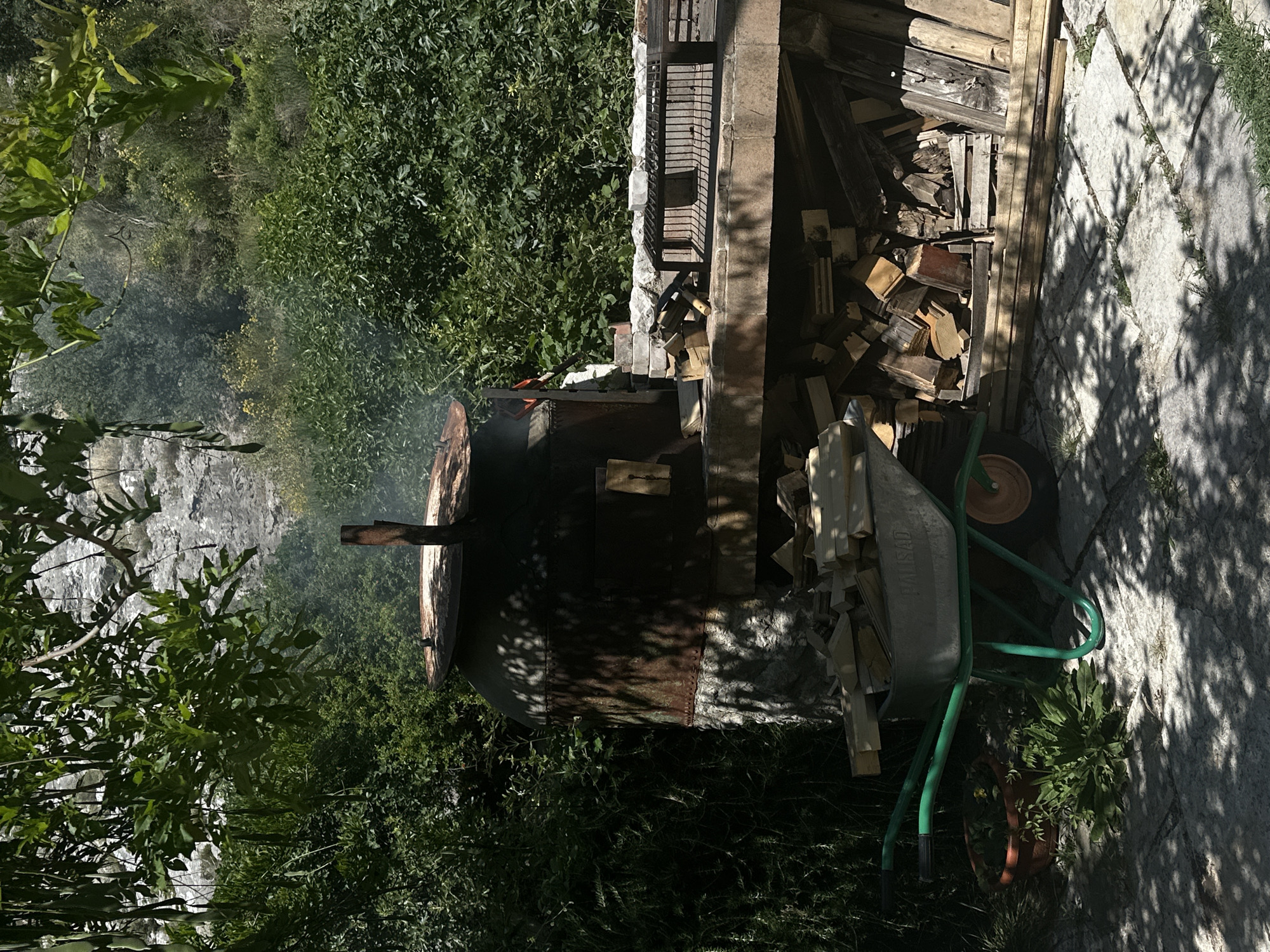 This outdoor scene in Bartula, Montenegro, features a rustic cooking or heating installation set against a lush, vegetated hillside under bright sunlight.
In the center-left, a large, dark, cylindrical metal structure, exhibiting significant rust and held together by rivets or seams, serves as an oven or smoker. It rests on a rough-hewn stone base and has a rusty metal chimney pipe protruding from its slightly conical top. Smoke is visibly rising from the top opening. Dappled shadows from overhead foliage pattern its surface and the stone base.
To the right of the oven, a sturdy, light-colored stone or brick counter provides a workspace. On this counter, a rectangular black metal grill grate is positioned. Smaller pieces of wood are scattered on the counter surface near the oven. Beneath the counter, a substantial quantity of cut firewood and various pieces of lumber are neatly stacked.
In the lower-left foreground, a bright green metal wheelbarrow, equipped with a single orange wheel, is positioned on a paved surface. It contains a load of split firewood. Adjacent to the wheelbarrow, a brown clay pot holds a small plant. The ground in the foreground is paved with light-colored, rectangular stone slabs, with grass visible in the gaps, and covered by strong, irregular shadows cast by overhead tree leaves.
The background is dominated by dense green foliage, comprising various bushes and trees. Beyond the immediate vegetation, a large, light-grey, rocky cliff face or steep hillside is partially visible through the trees. The scene is illuminated by strong natural sunlight, creating high contrast and distinct shadows. No people are present in the image.