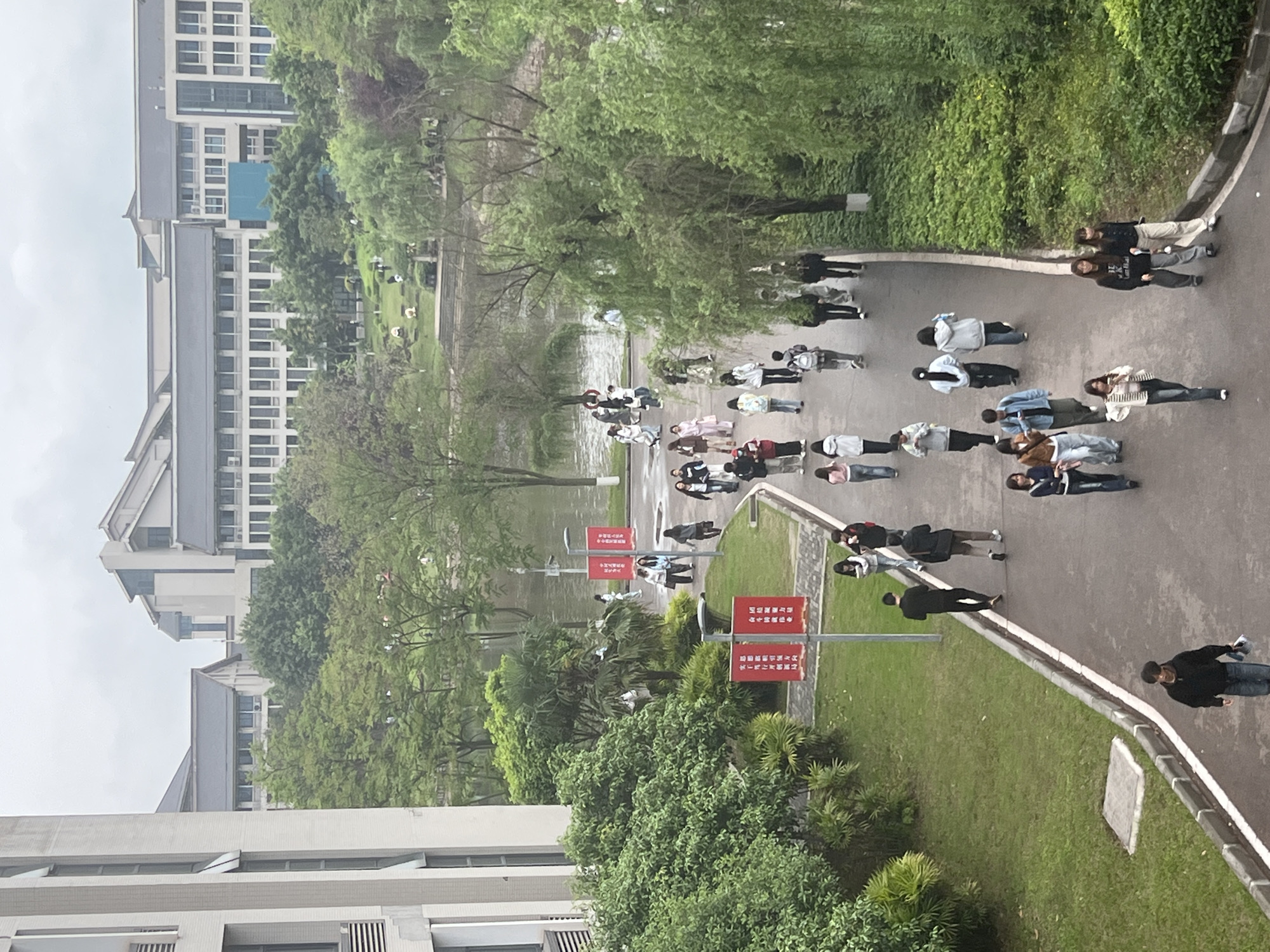 The image depicts an outdoor scene in Jiangjin, China, likely on a university campus. A group of people, mostly young adults, are walking or standing on a paved path. The path runs adjacent to a body of water and is lined with trees and green spaces. Several red banners with Chinese writing are visible along the path. Buildings with multiple floors are present in the background. The sky is overcast.