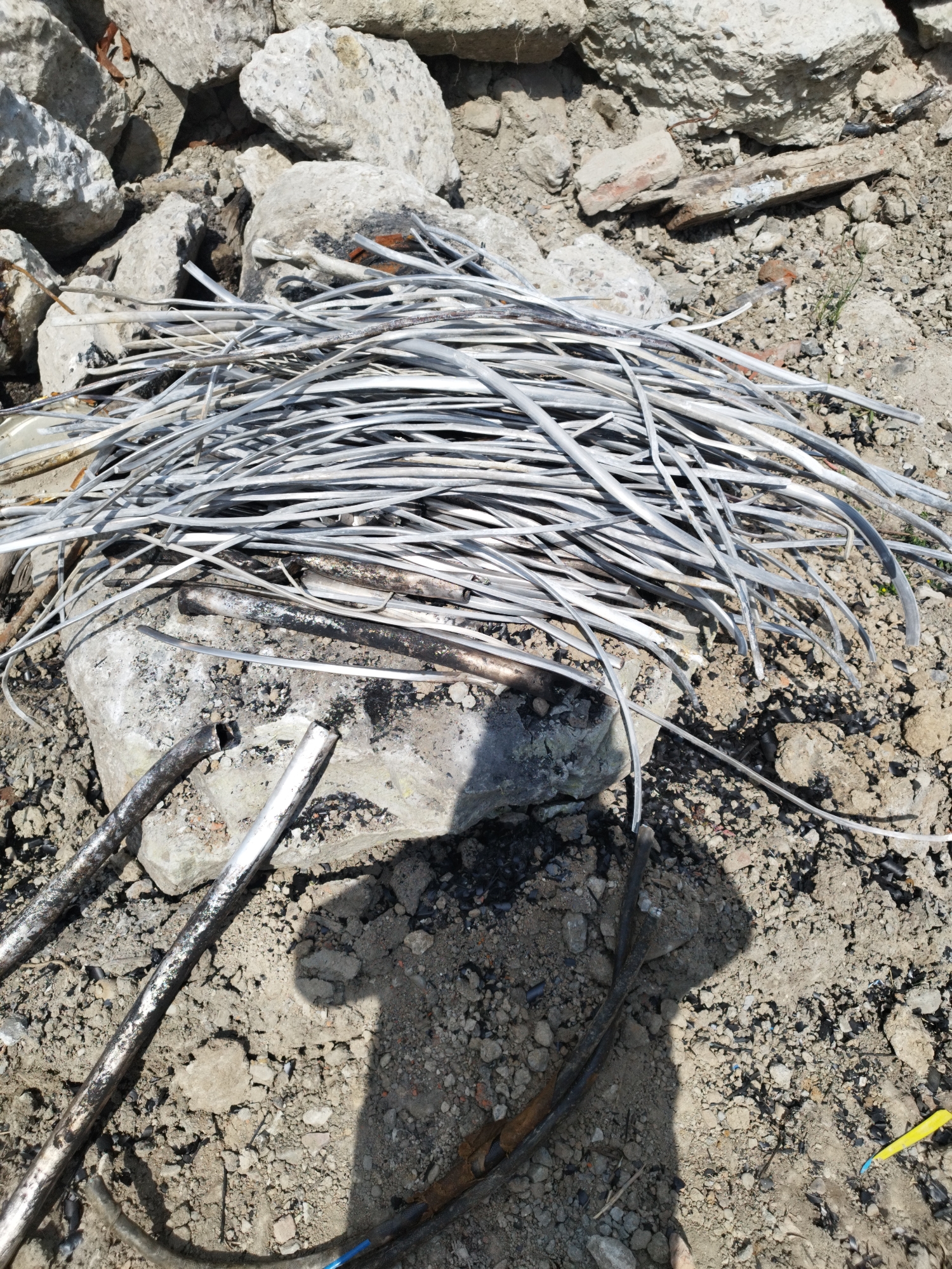 This media file depicts a pile of discarded aluminum wires and debris on the ground in Rozdil, Ukraine. The scene appears to be an outdoor environment, likely a construction or demolition site, characterized by rubble, broken concrete, and loose soil.
The central subject is a tangled mass of thin, silvery aluminum wires, some of which are bent and curved, indicating they have been used and discarded. Beneath the wires, a large, grey, broken concrete slab serves as a base, with more debris scattered around it. Two thicker, metallic rods with a weathered, reflective surface are also visible in the foreground, resting on the concrete. In the lower portion of the image, dark, pliable cables, possibly electrical or communication wires, are visible amongst the dirt and stones. A small, bright yellow object, possibly a piece of discarded plastic or tape, is also present in the lower right corner.
The lighting suggests it is daytime, with sunlight casting shadows, including a prominent shadow of a person on the ground, indicating the presence of a photographer or someone nearby. The overall impression is one of neglect and a post-construction or demolition aftermath. No specific activity or event is actively taking place; the image captures a static scene of discarded materials. No text is visible in the media file.