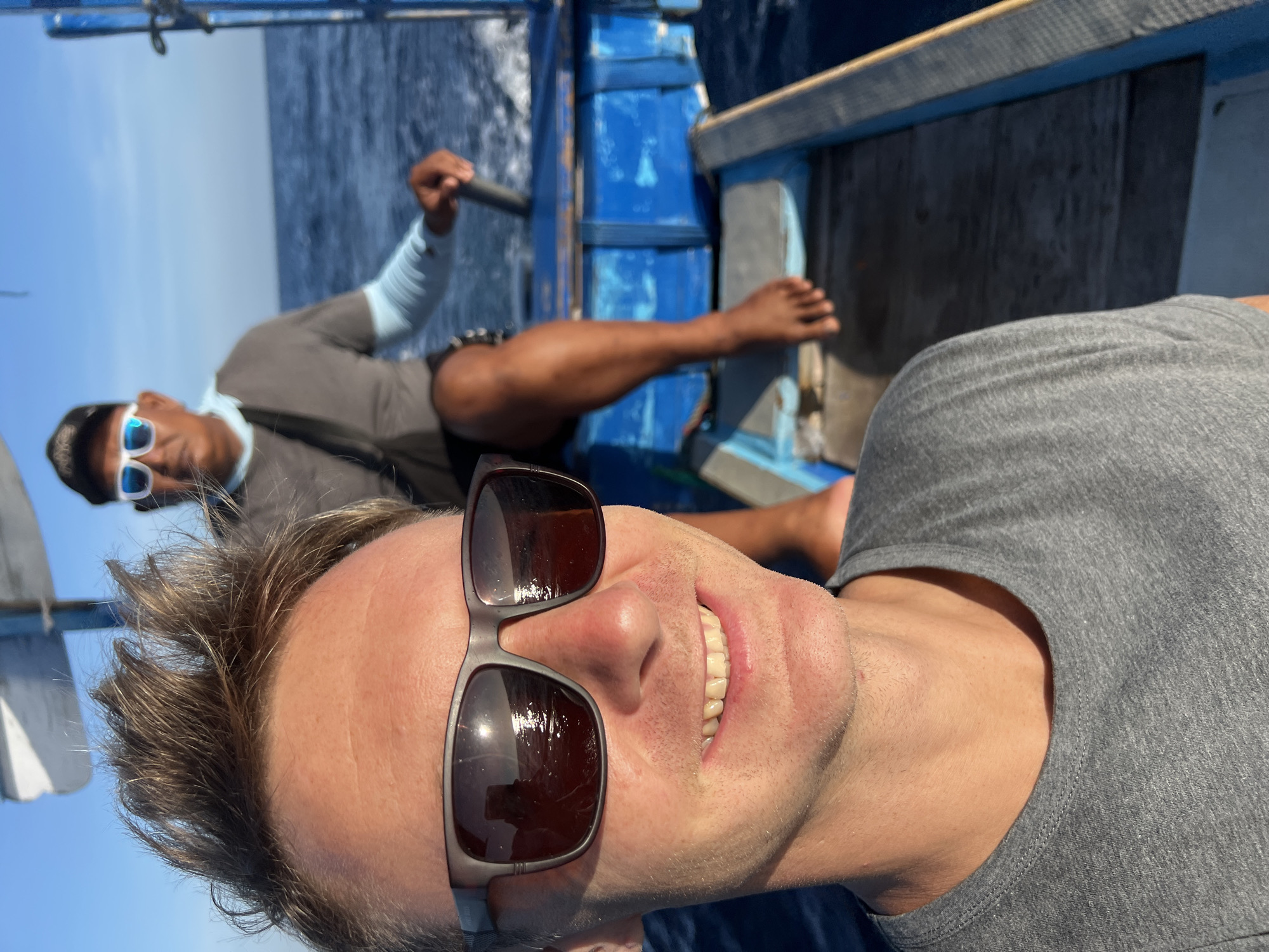 A selfie photograph taken on a boat. A light-skinned man with short brown hair, wearing sunglasses and a gray t-shirt, smiles at the camera. In the background, a dark-skinned man wearing sunglasses and a gray shirt with light blue sleeves rows the boat with a paddle. The boat is blue. The setting appears to be the sea. The photo was taken in Seririt, Indonesia.