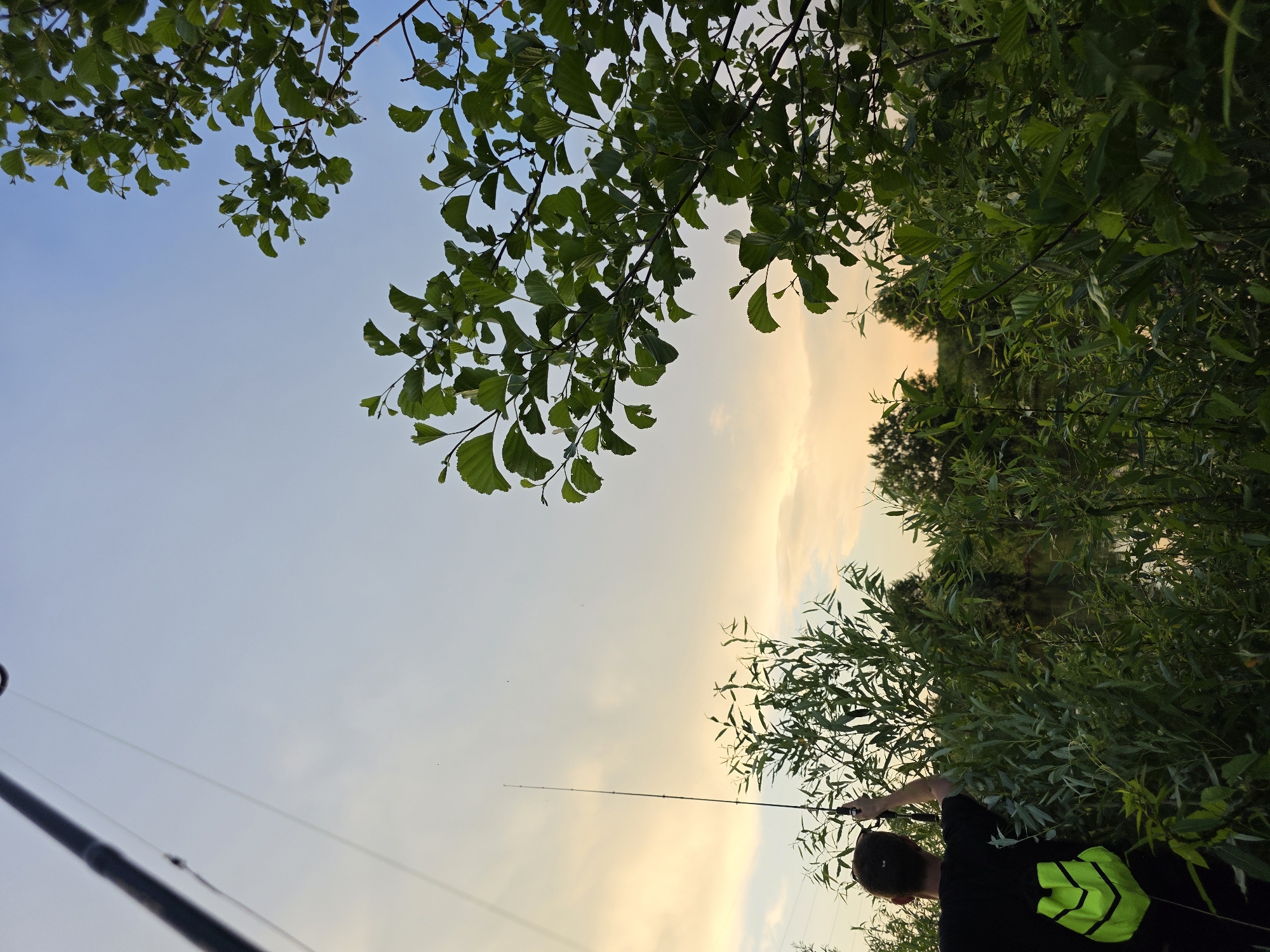 A low-angle, medium shot captures a person fishing on the shore of a body of water in Togashevo, Russia. The person, viewed from behind, wears a black t-shirt and a neon green backpack. They hold a fishing rod and are partially obscured by dense green foliage. The sky above transitions from a pale blue near the top to a warm, soft orange and yellow near the horizon, indicating either sunrise or sunset. Lush green trees and bushes dominate the foreground and mid-ground, with the water visible through the vegetation. The overall scene suggests a peaceful, natural setting.
